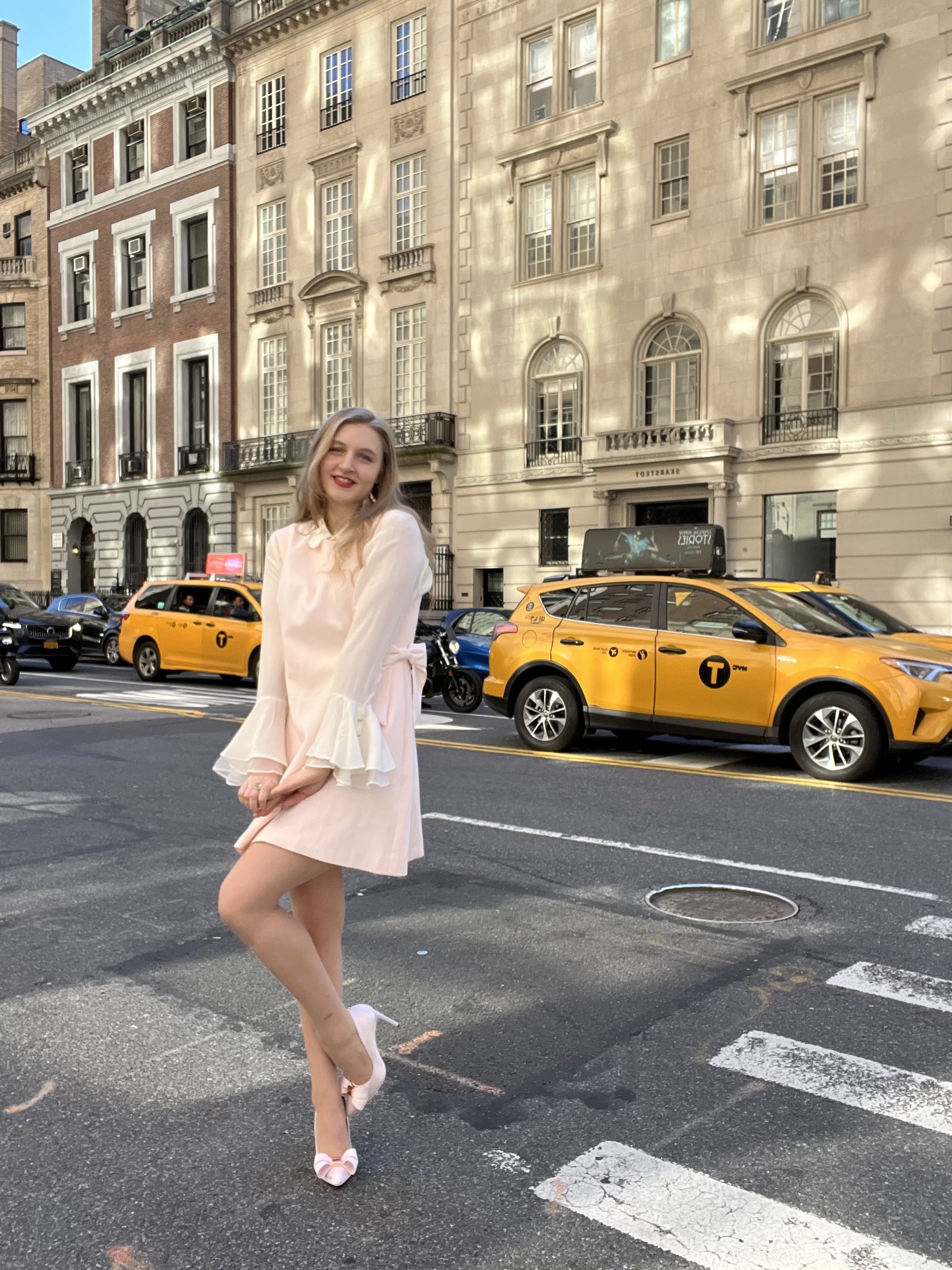 Jacqui de Jong wears a silk white shirt with bell sleeves with a light pink minidress. She wears light pink bow heels.  