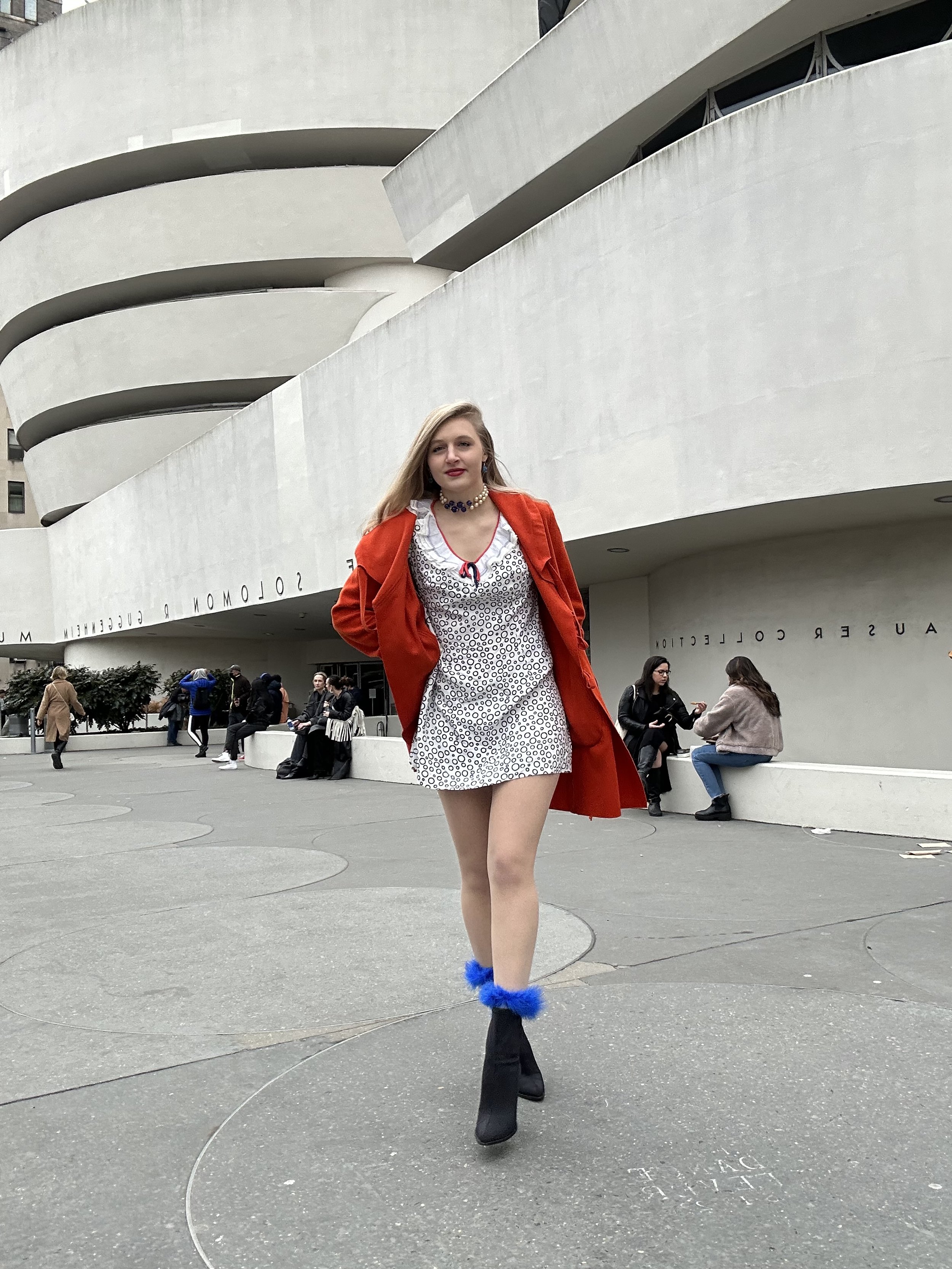 Jacqui de Jong wears a 1960s blue and white  polka dot dress with a blue and white choker necklace. She also wears a large orange cashmere coat, blue feather socks and black boots.  