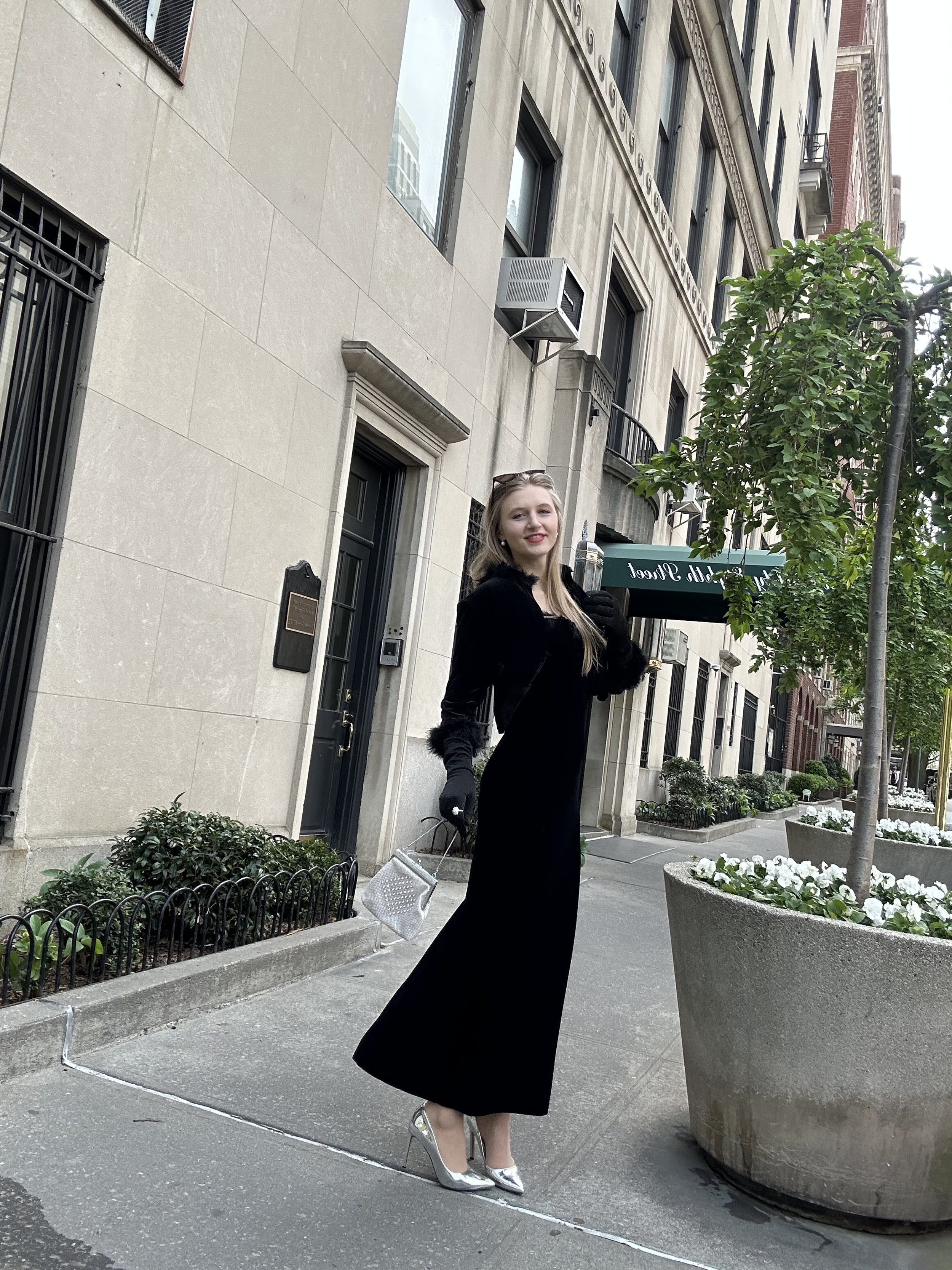 Jacqui de Jong wears a long velvet evening gown from the 1990s, with a matching cardigan with fluffy feather trim on the collar and sleeves. She also wears long black gloves, Prada sunglasses and silver heels. 