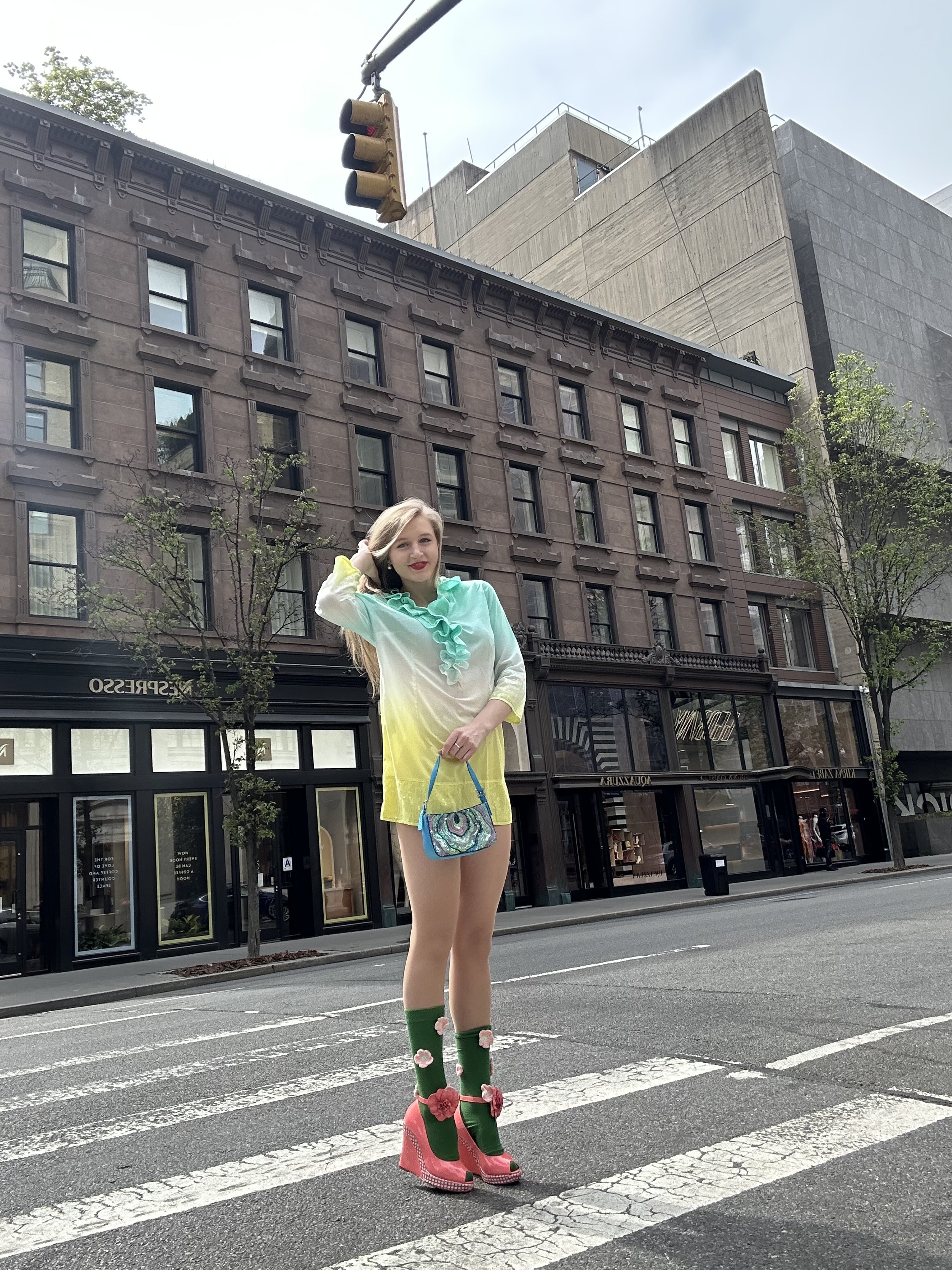 Jacqui de Jong wears a yellow, white and aqua dress with a ruffled collar from the early 2000s. She pairs the look with green and pink knee-high flower socks, and bright pink wedge sandals. She accessorizes the look with a blue sparkly sequin handbag