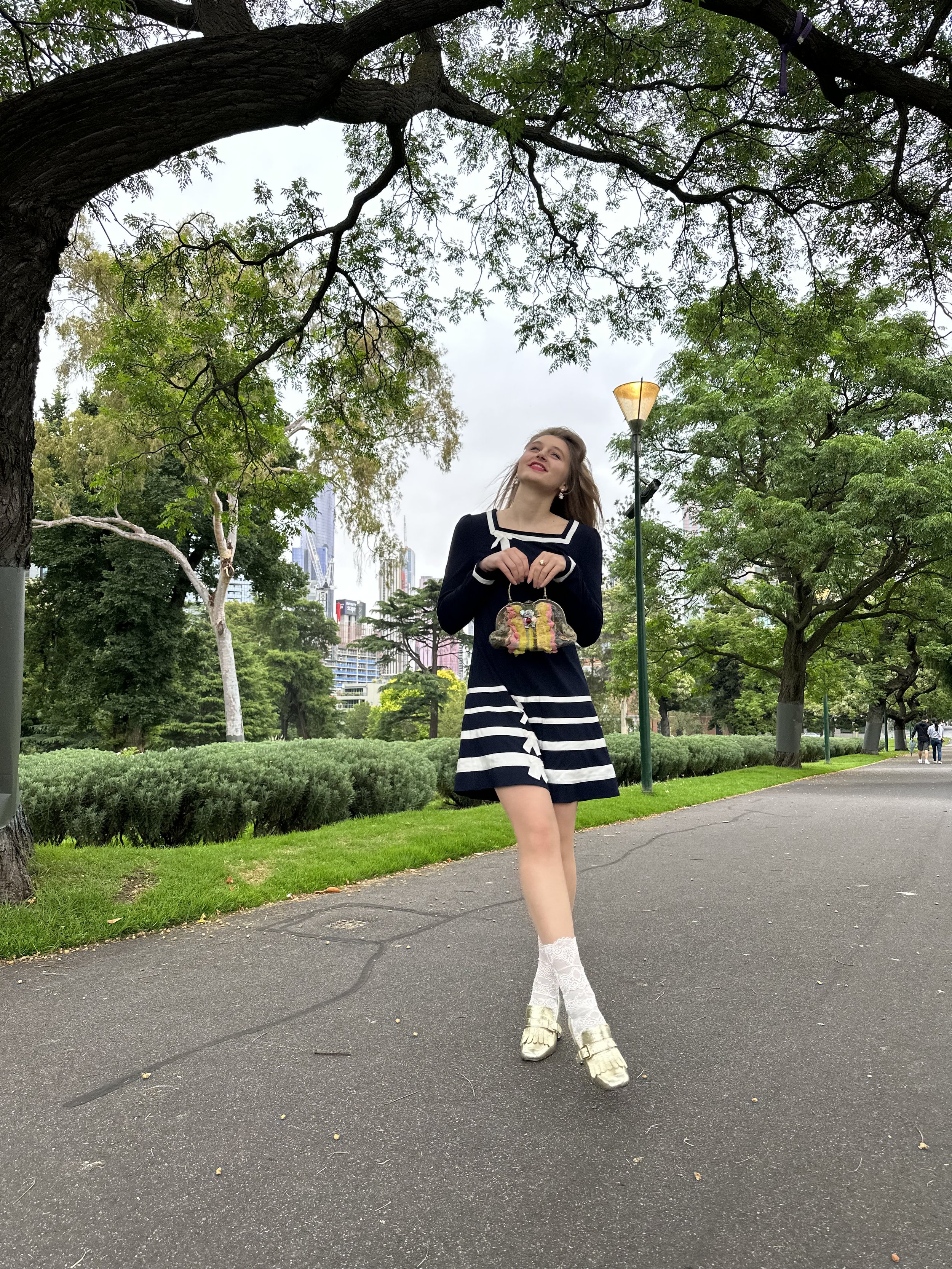 Jacqui de Jong wears a navy blue dress with a square collar and white stripes on the skirt hem, with bow detail. She also wears white lace socks, gold leather shoes and has a rainbow vintage purse.