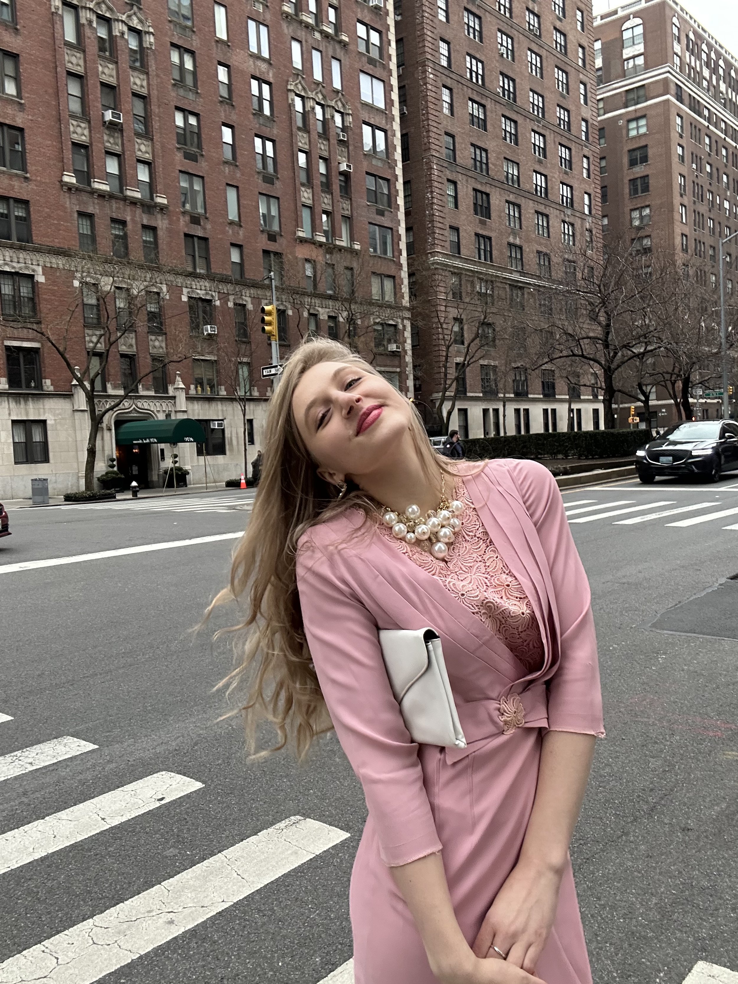 Jacqui de Jong wears a vintage light pink dress with a bow at the waist and pink lace on the bodice. She wears a large pearl necklace and holds a vintage white purse.