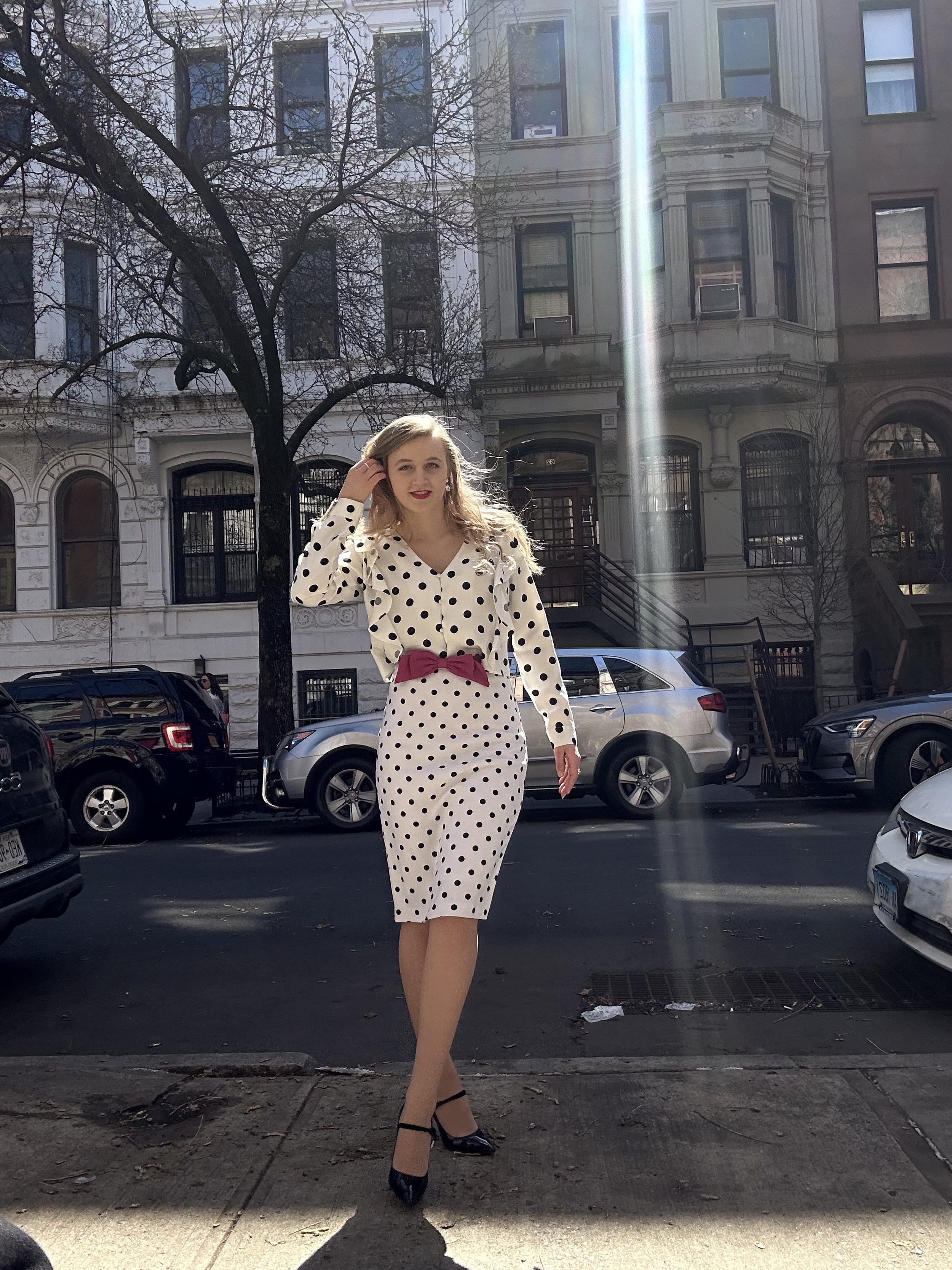 Jacqui de Jong wears a polka dot black and white pencil skirt with a polka dot black and white long-sleeved shirt with a V-neck collar and single edge ruffles on each front panel. Look is accessorized with a pink bow belt and black stiletto shoes. 