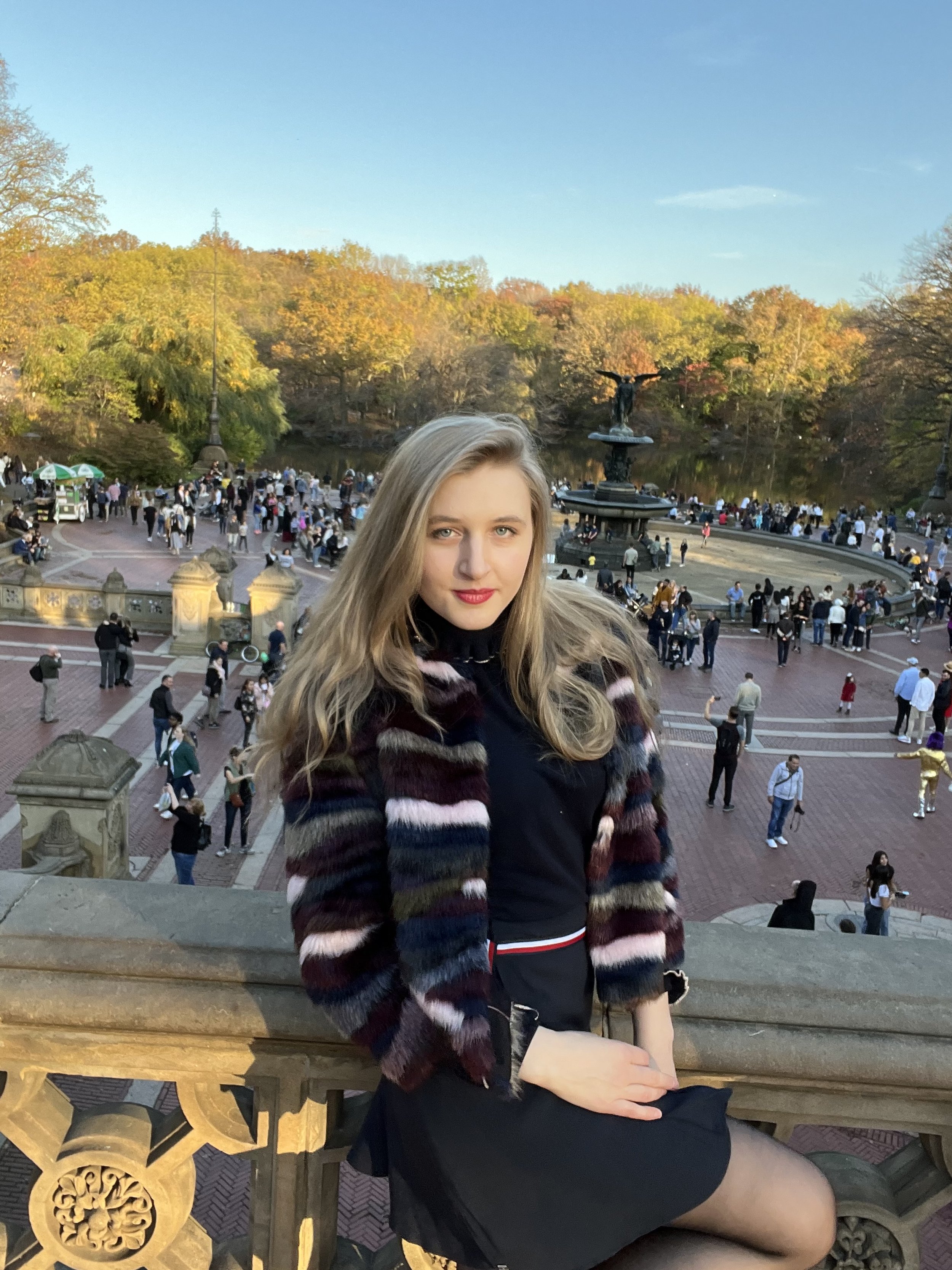Jacqui de Jong wears a navy blue skirt with a black, white and red tie around the waist. She also wears a brown, white, blue, green, black and cream fur coat, and a navy blue turtleneck shirt. 