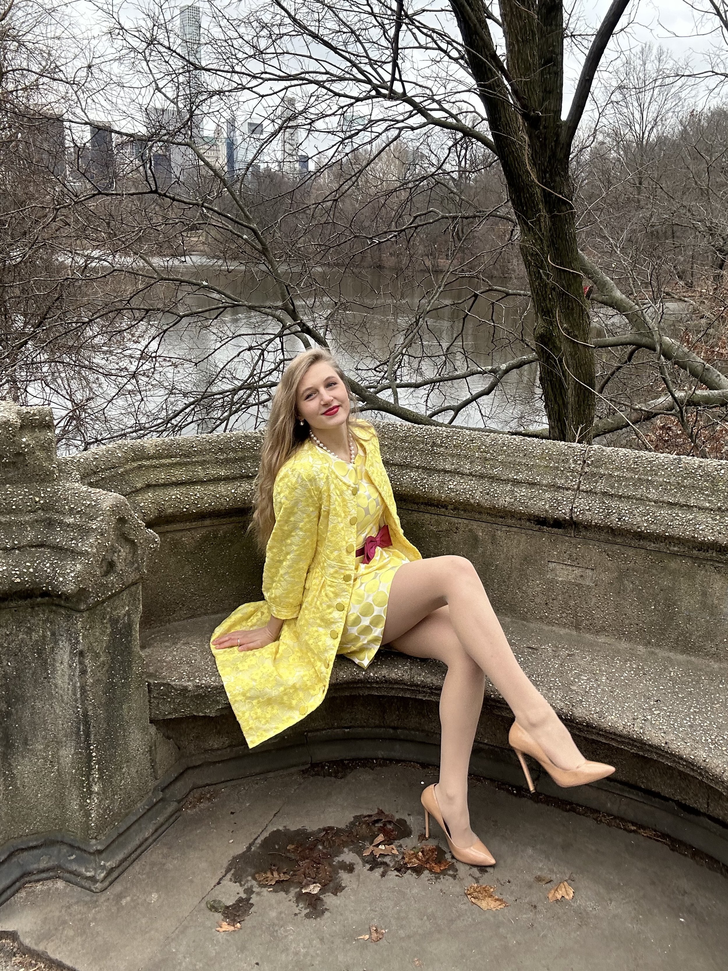 Jacqui de Jong wears a lemon polka dot dress with a large yellow lace coat, accessorized with a pink bow belt around the waist. She completes the look with nude shoes. 