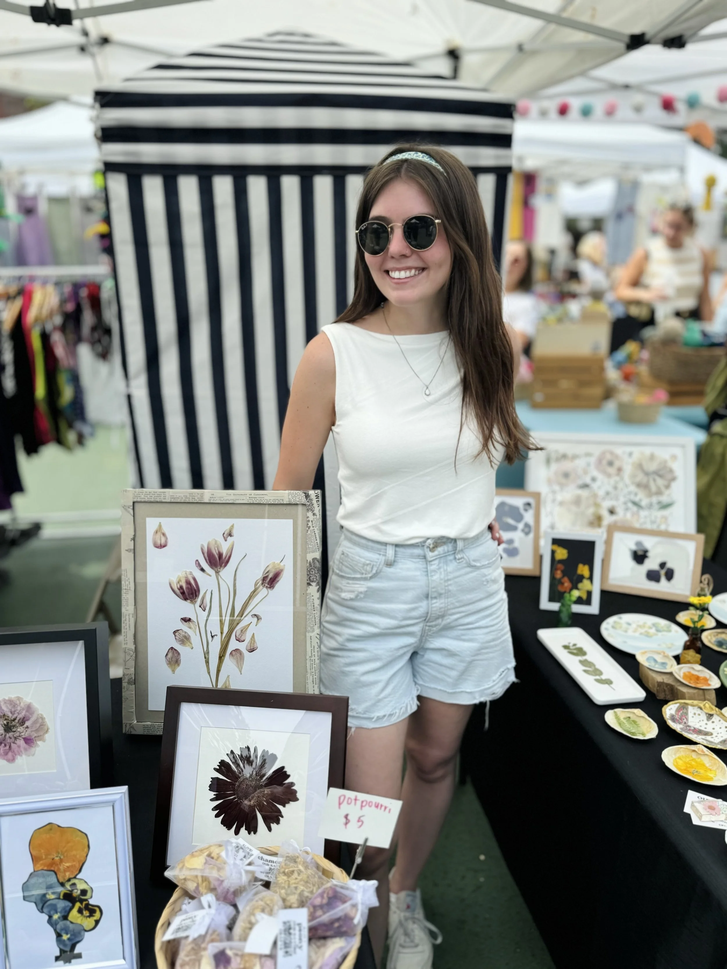 A woman with long brown hair, sunglasses, a white sleeveless top, and light denim shorts standing behind a booth at a craft fair, surrounded by artwork and small decorative products.