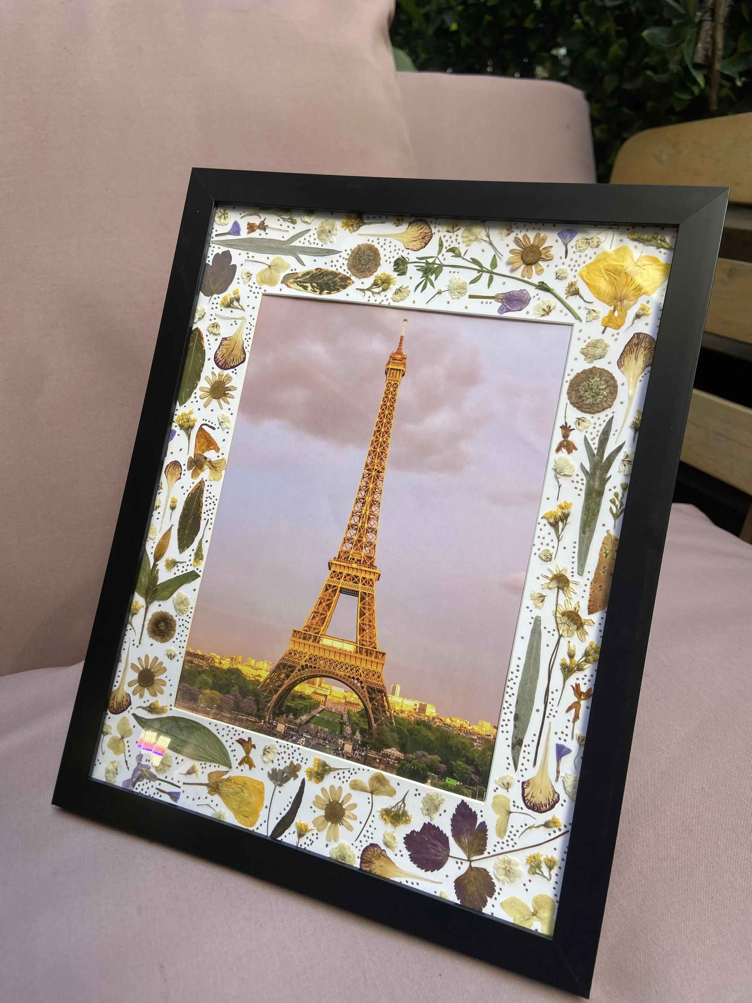 Framed photo of the Eiffel Tower with a decorative border of pressed flowers and leaves on a pink fabric surface.