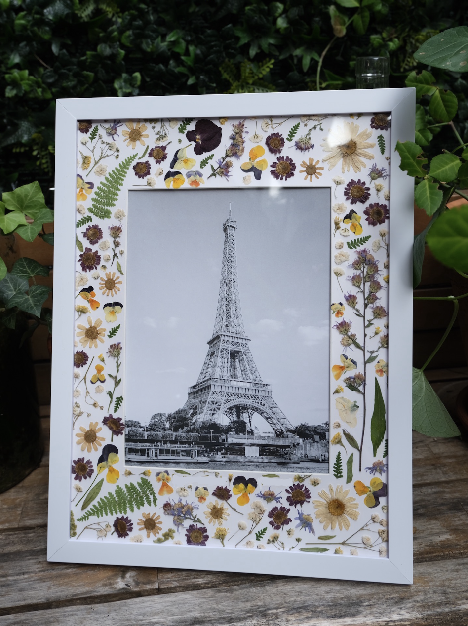 Framed black and white photo of the Eiffel Tower decorated with pressed flowers and leaves, placed on a wooden surface with green leafy plants in the background.