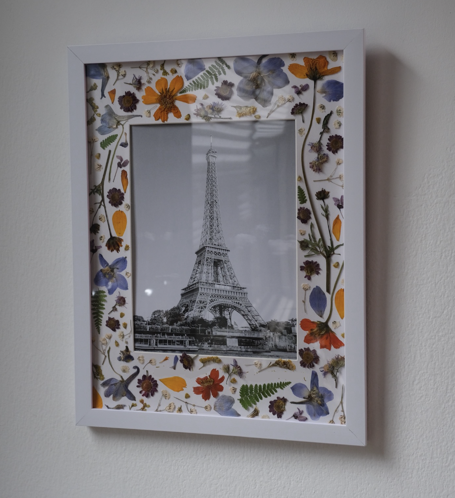 Framed black and white photo of the Eiffel Tower surrounded by a white frame decorated with pressed colorful flowers and green leaves.