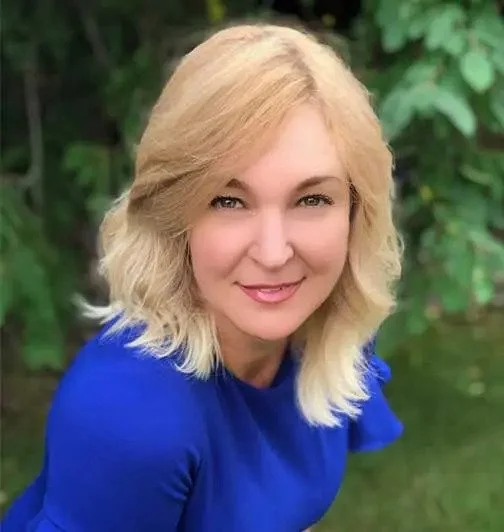 A woman with blonde wavy hair smiling outdoors in front of green foliage, wearing a blue top.