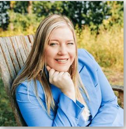 Portrait of a smiling woman with blonde hair, wearing a blue blazer, sitting outdoors on a wooden bench with trees and grass in the background.