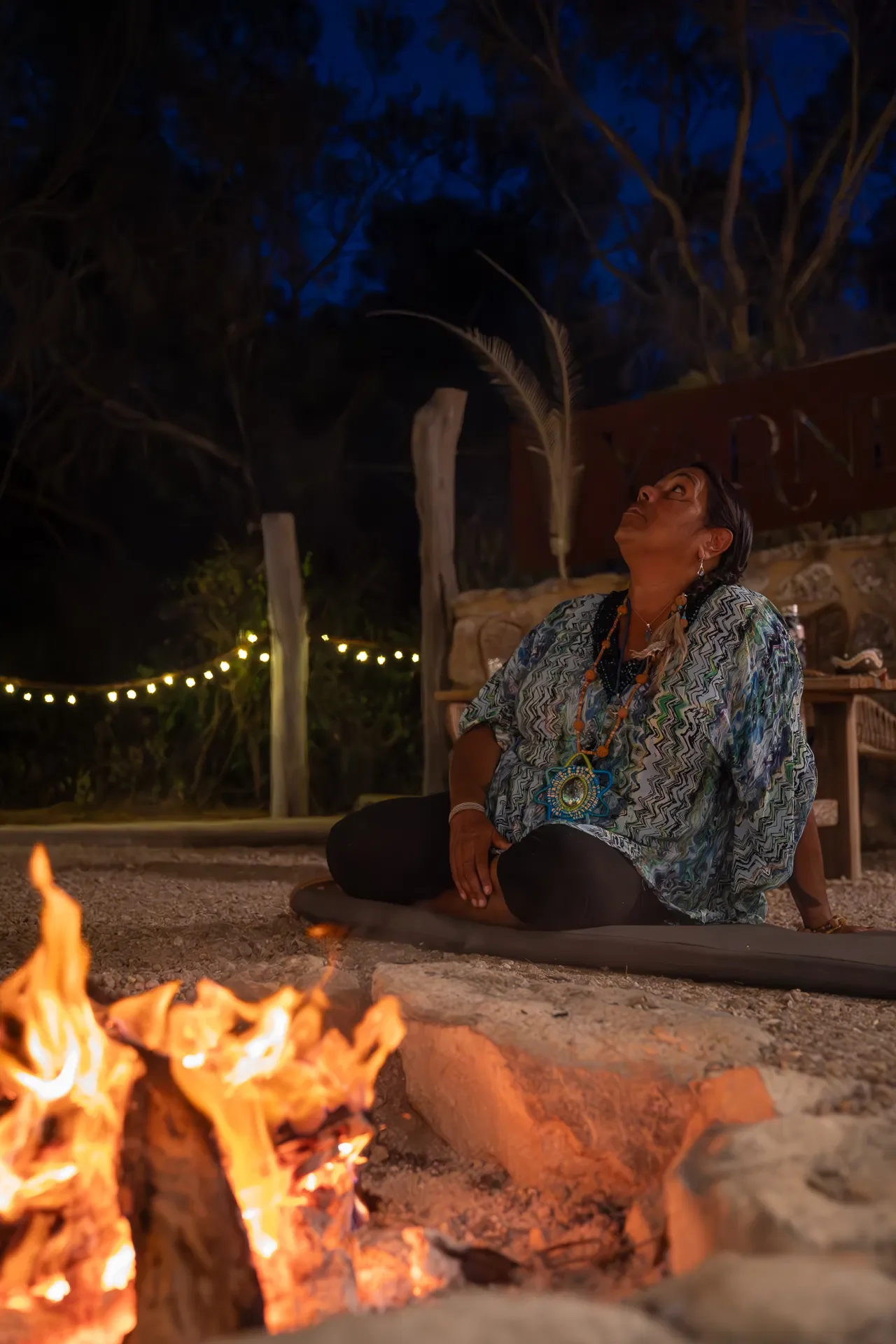 A woman sitting cross-legged on a mat near a campfire during the evening or night, with trees and string lights in the background, looking upward.