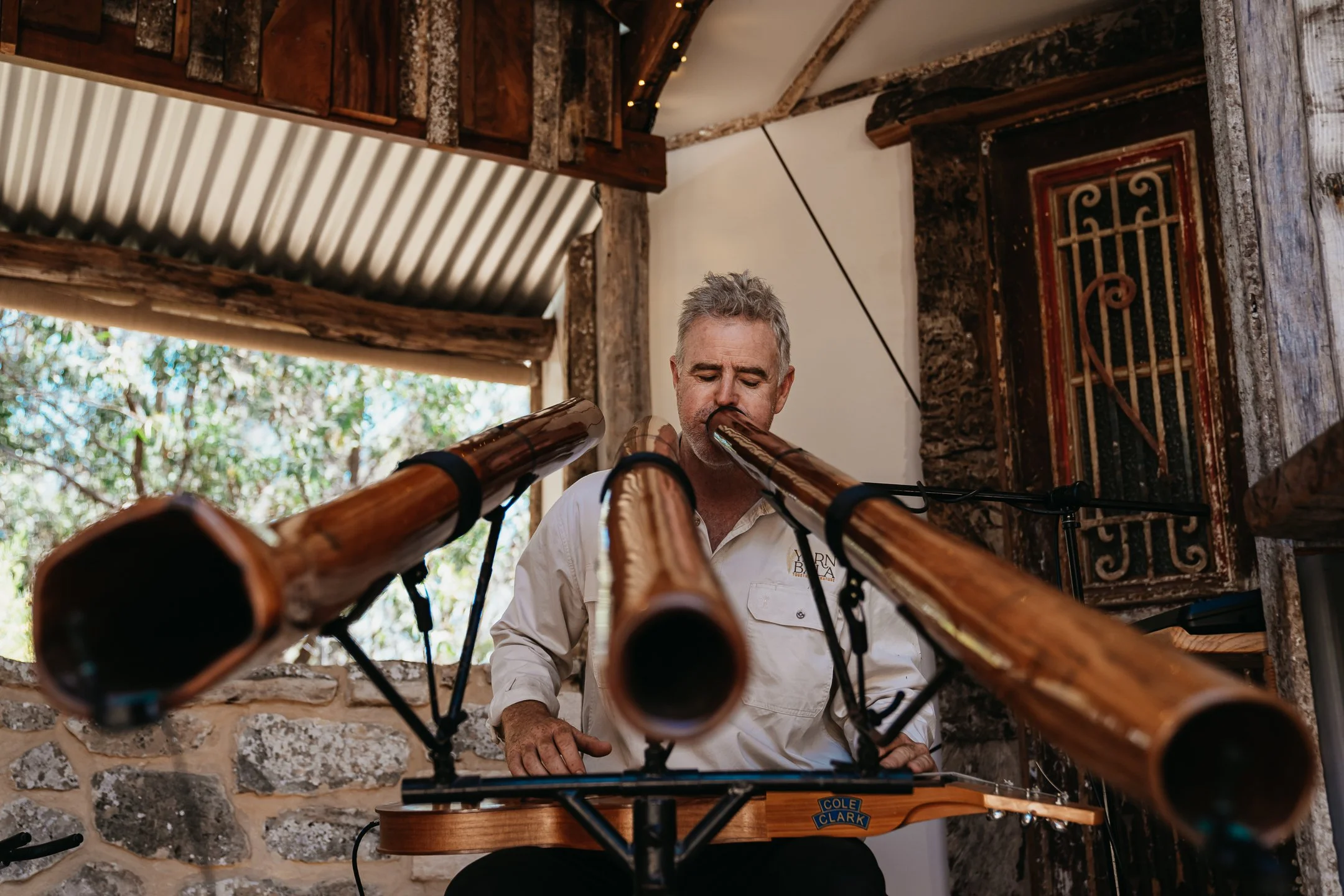 A man playing a long wooden flute or didgeridoo indoors with a rustic wooden and stone background.