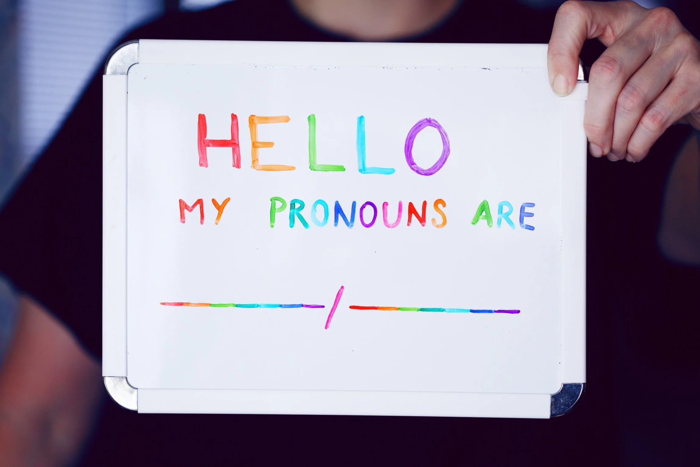 a white person is holding up a dry erase board with HELLO MY PRONOUNS ARE ____ / ____ written in rainbow colors