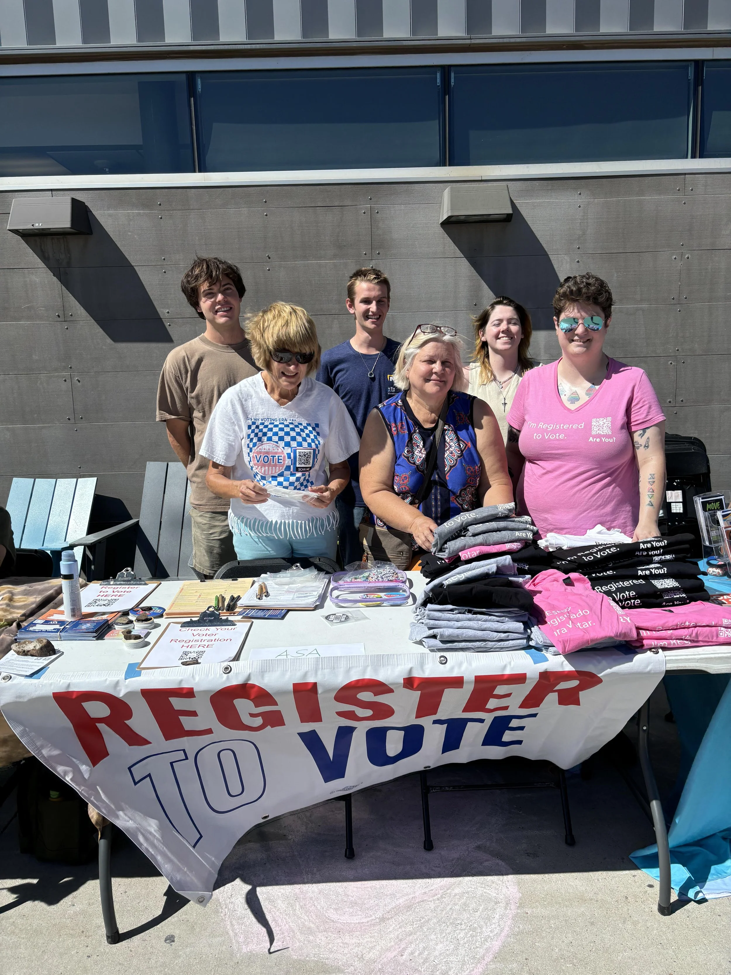 6 people gathered behind a table with papers and t-shirts on it and a banner that says "Register to Vote"
