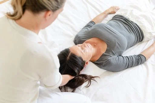 Eine Frau liegt auf einem Bett und eine Therapeutin, hält ihren Kopf sanft. Es ist eine Shiatsubehandlung um das Nervensystem zu entspannen.