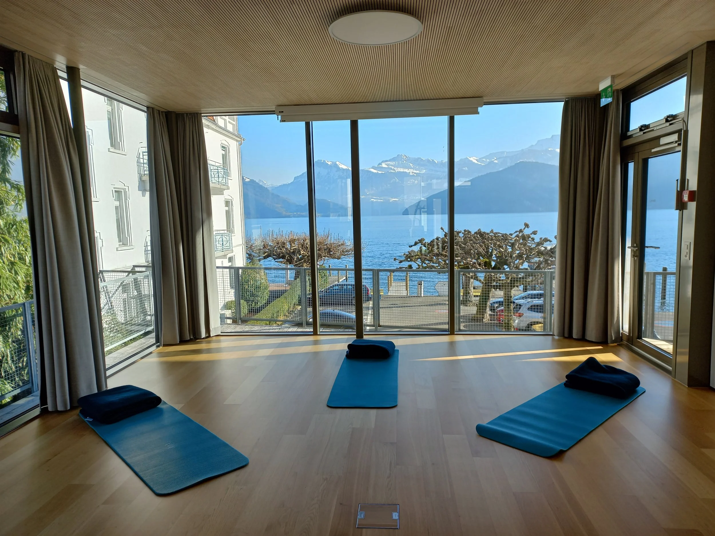 Innenraum eines Raums mit großen Fenstern, die einen Blick auf Berge, See und Himmel bieten. Auf dem Boden liegen drei Yogamatten mit gefalteten Handtüchern darauf, bereit für eine Yoga- oder Meditationssitzung.