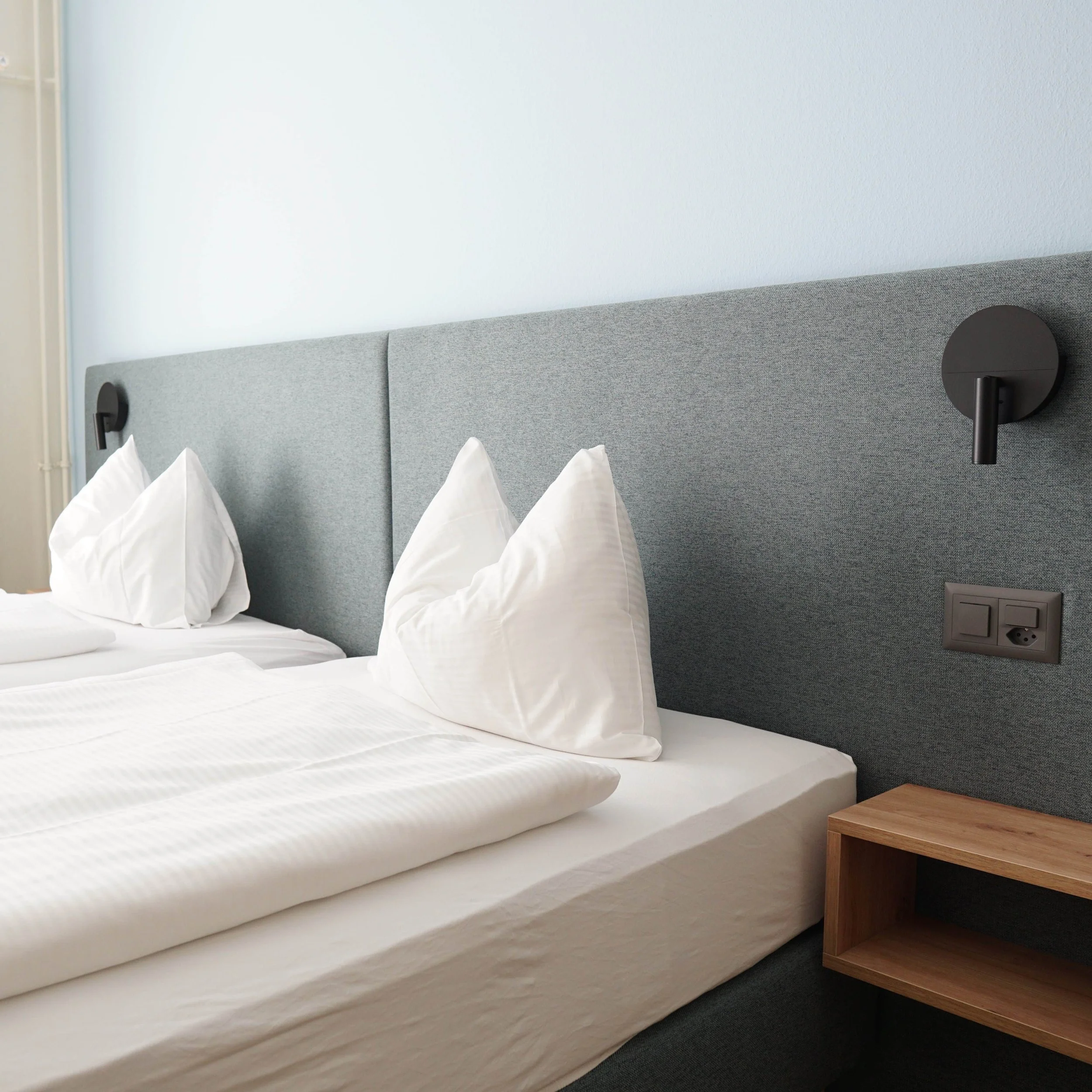 Bett mit weißen Kissen und Bettwäsche in einem modernen Hotelzimmer, grauer gepolsterter Kopfteil, Wandlampe und Steckdose auf der rechten Seite.