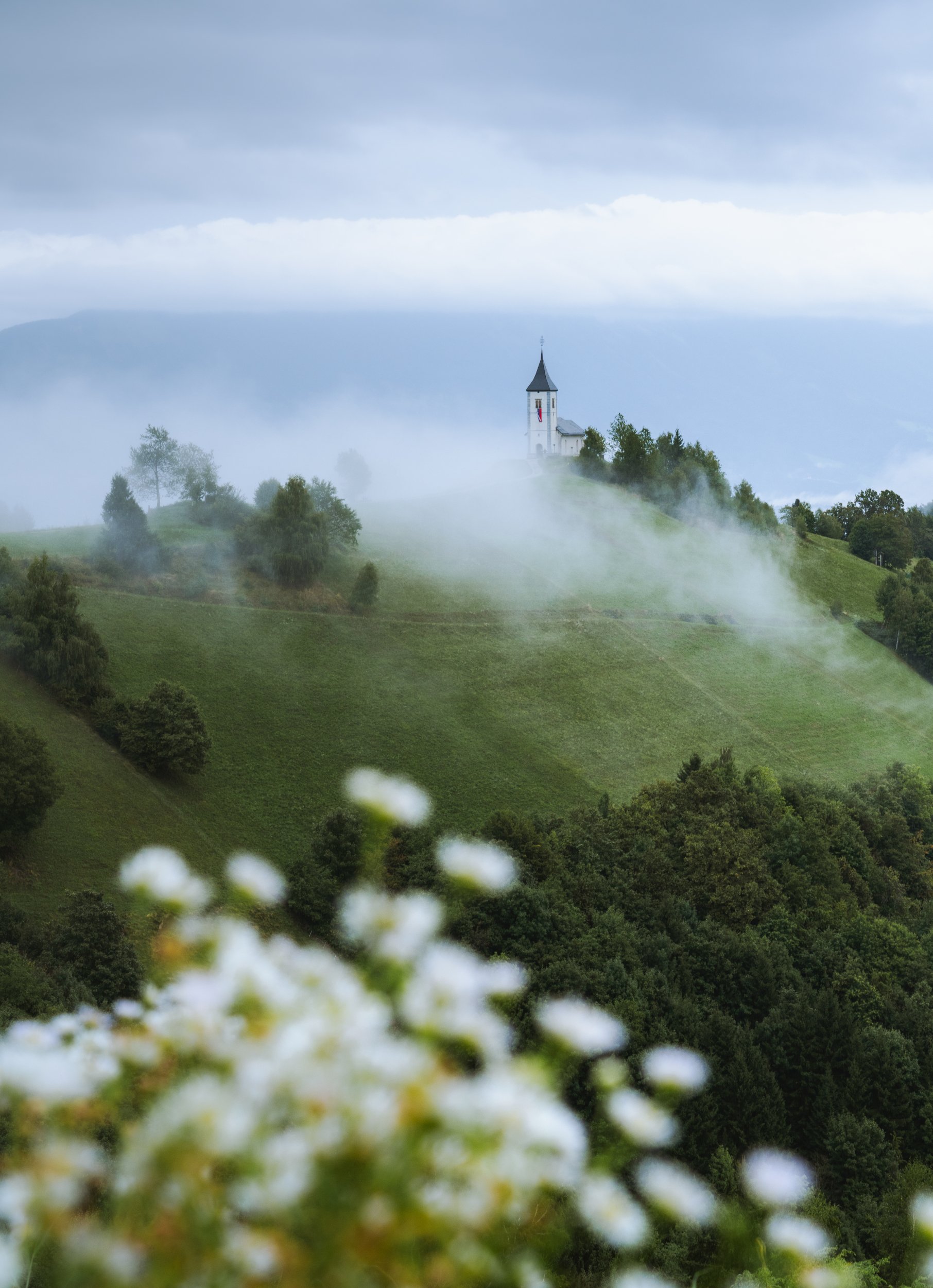 A small white church with a pointed steeple is situated on a hill surrounded by green trees and grass, with fog rolling over the landscape.