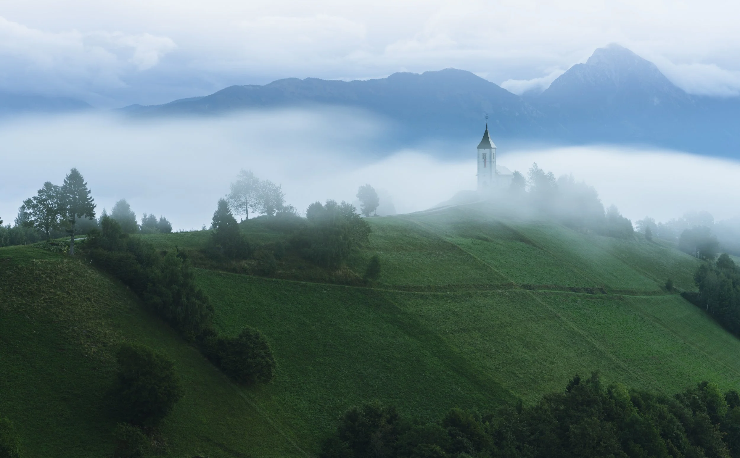 A church on a green hill surrounded by trees with a mountain range in the background, fog covering the landscape.