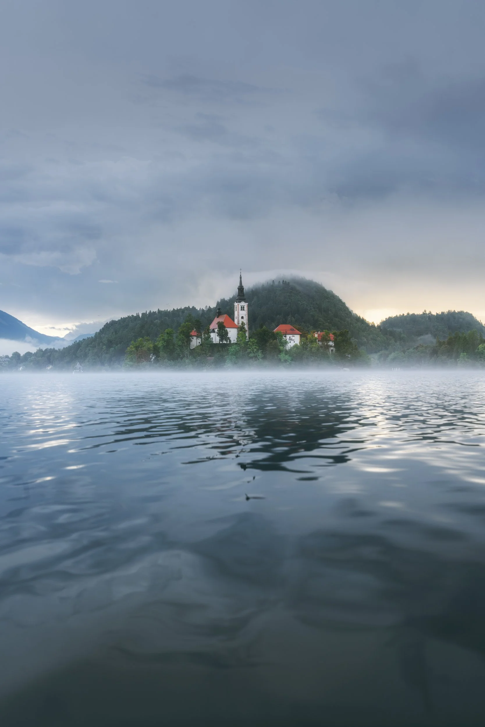 A scenic view of a small church with a tall steeple on an island surrounded by a misty lake, with forested hills and mountains in the background under a cloudy sky.