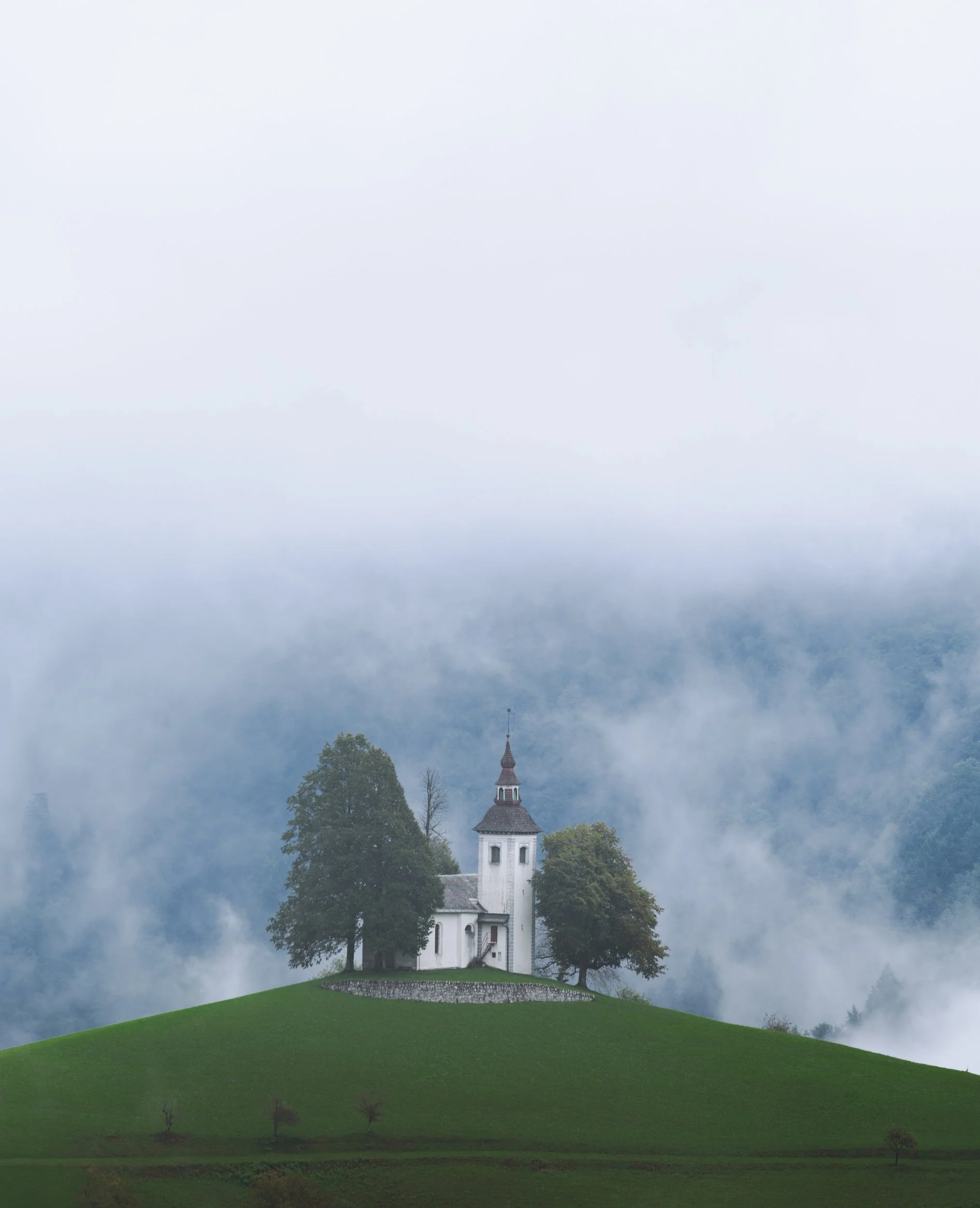A small white church with a steeple on a grassy hill, surrounded by trees and enveloped in fog, with mountains in the background.