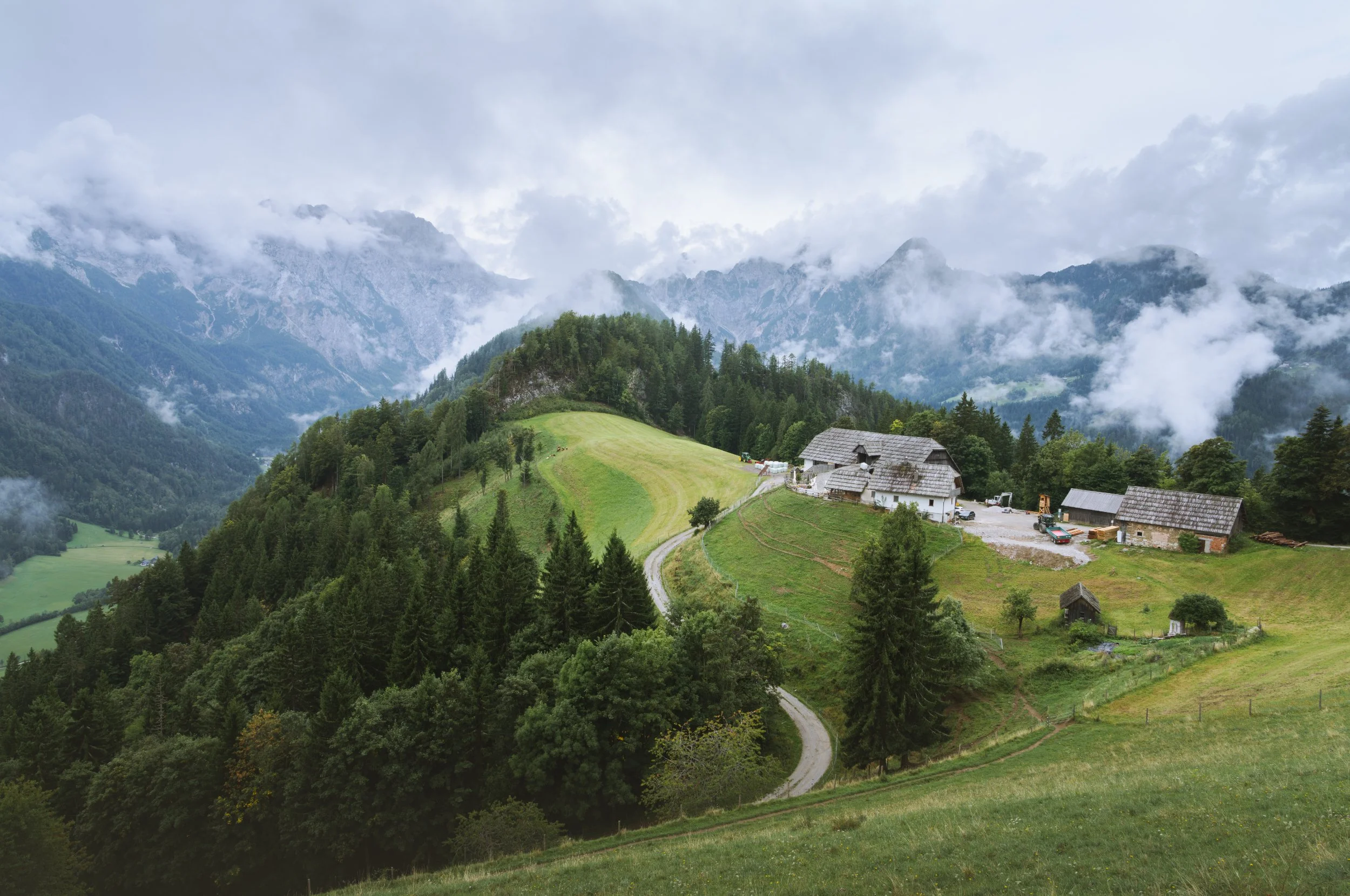 Green hillside with scattered trees and farm buildings, winding dirt path, mountains in the background shrouded in clouds and mist