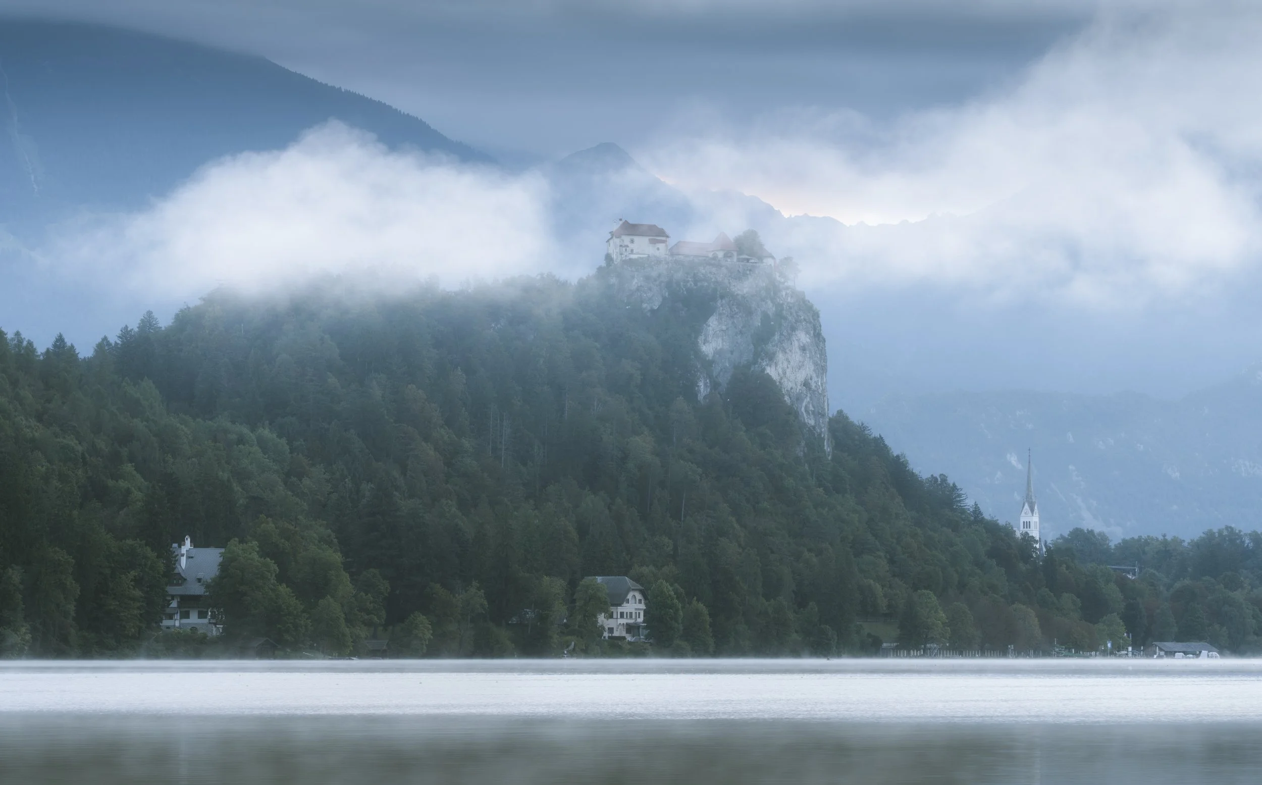 A foggy mountain landscape with a castle on a rocky cliff, surrounded by dense trees, with a lake and houses at the base, and a church steeple in the distance.