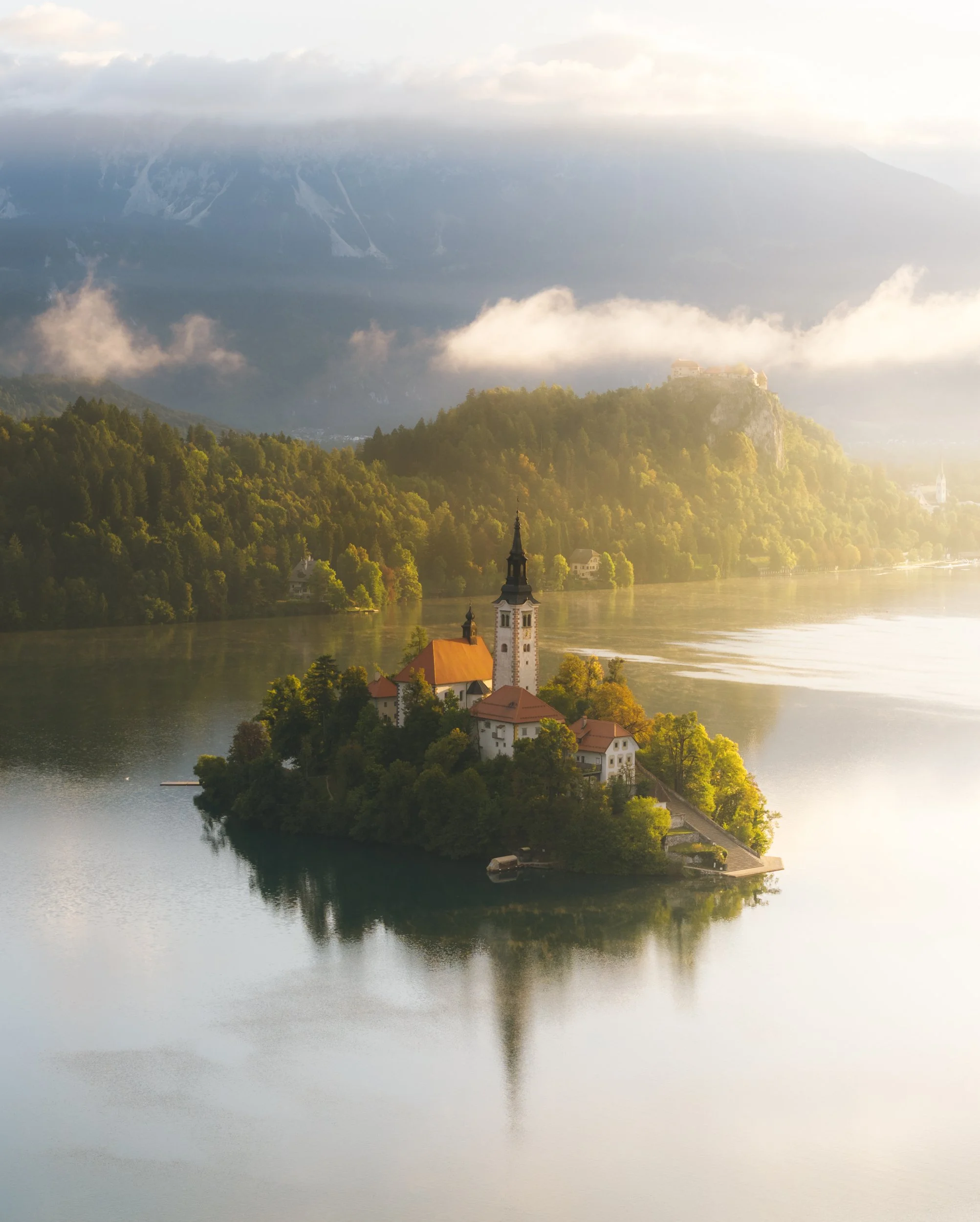 A small island with a church and other buildings, surrounded by calm water, trees, and mountains in the background, bathed in soft sunlight.
