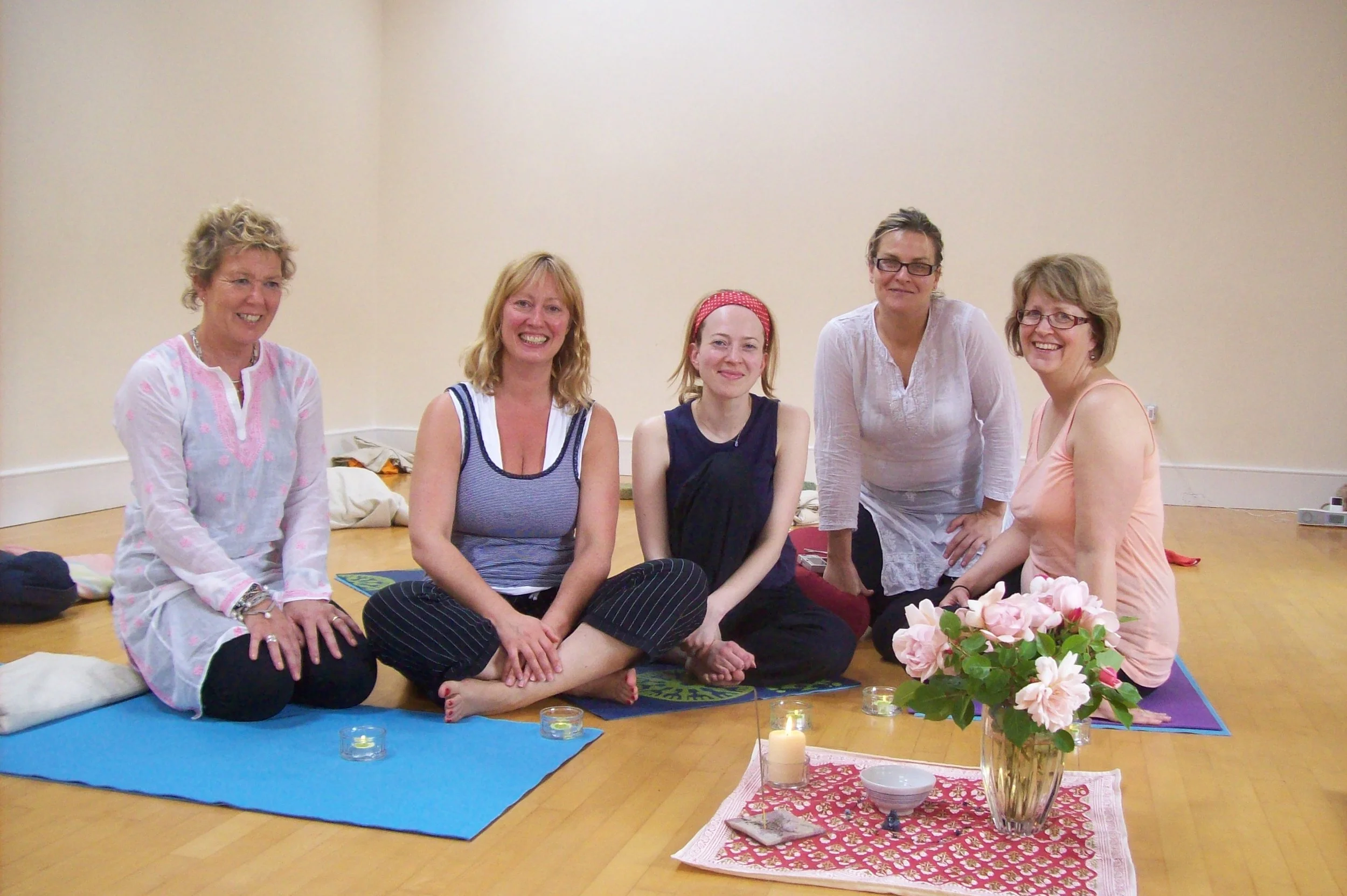 Five women sitting on yoga mats in a room, with a small altar featuring a lit candle, a bowl, and pink flowers in a vase in front of them.