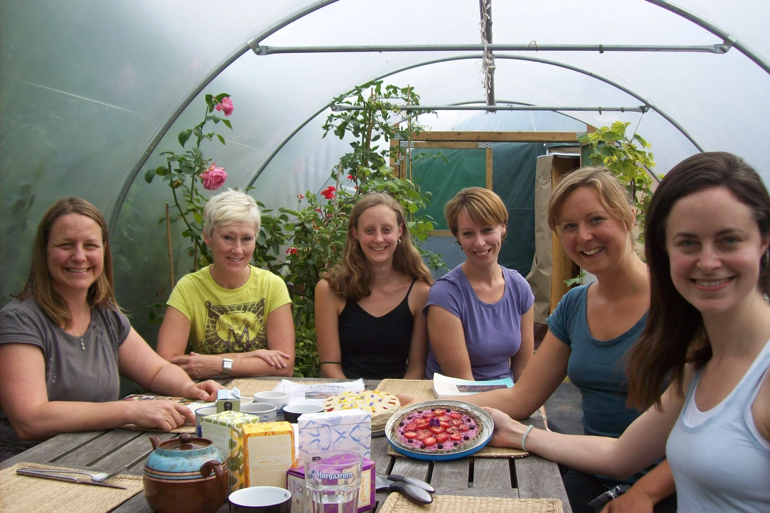 Six women sitting at a table inside a greenhouse, celebrating with cakes and drinks.