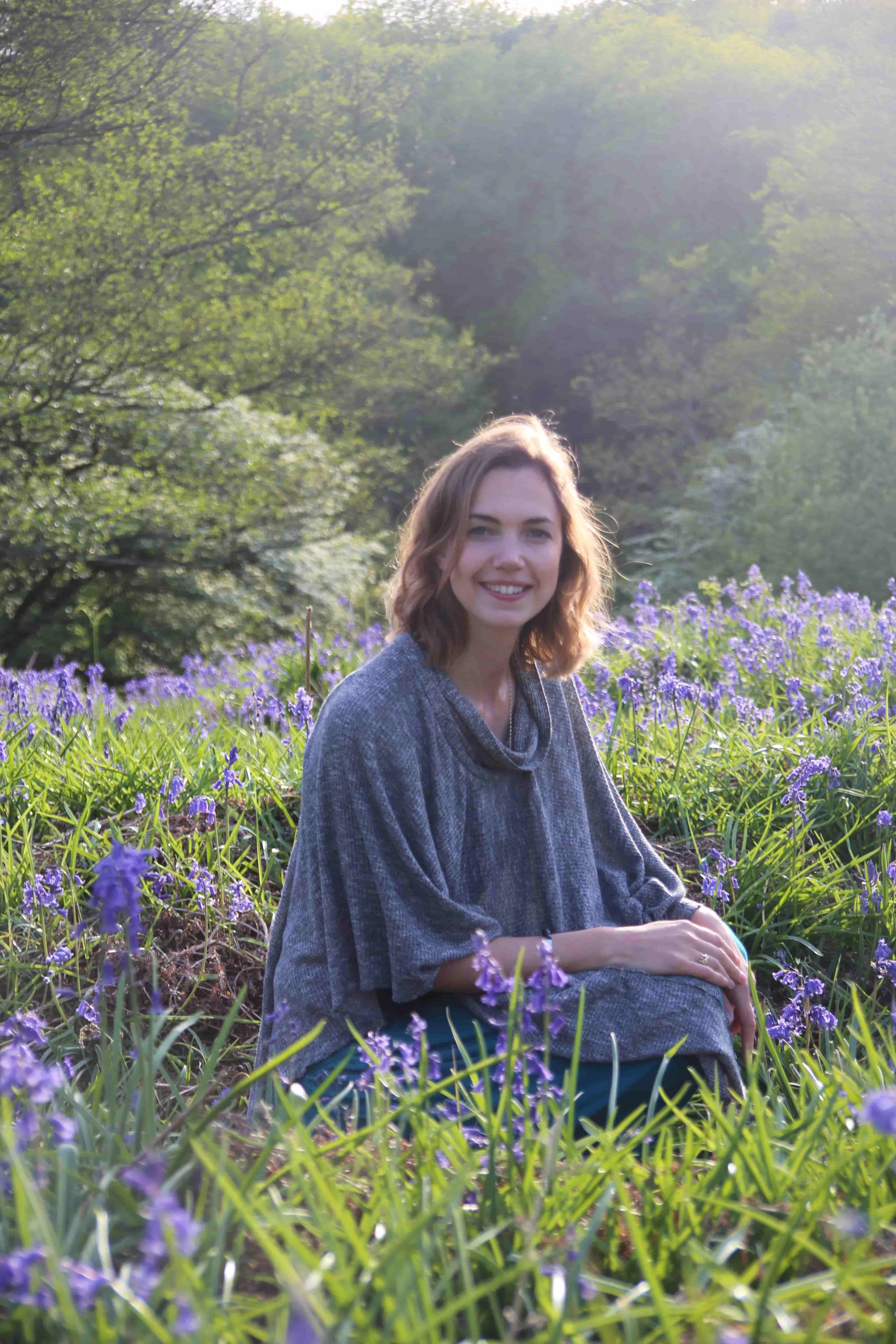 A young woman sitting in a field of purple flowers with trees and sunlight in the background.