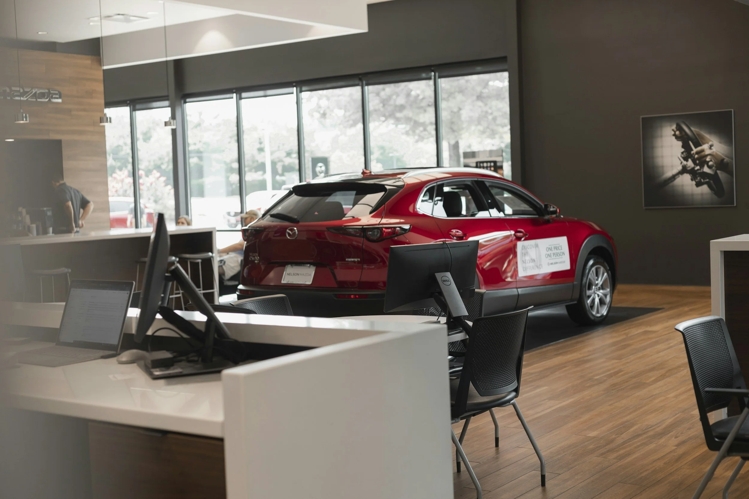 A red Mazda car displayed inside a modern car dealership showroom with desks, chairs, and computers visible in the foreground.