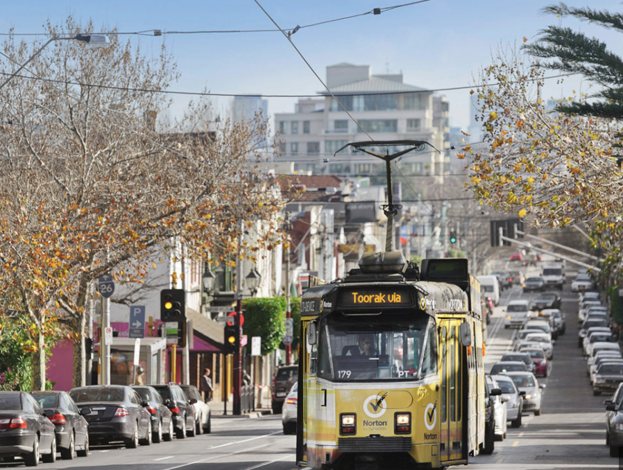 A yellow tram moving down a busy city street lined with parked cars, trees, and buildings, with some trees having autumn leaves.