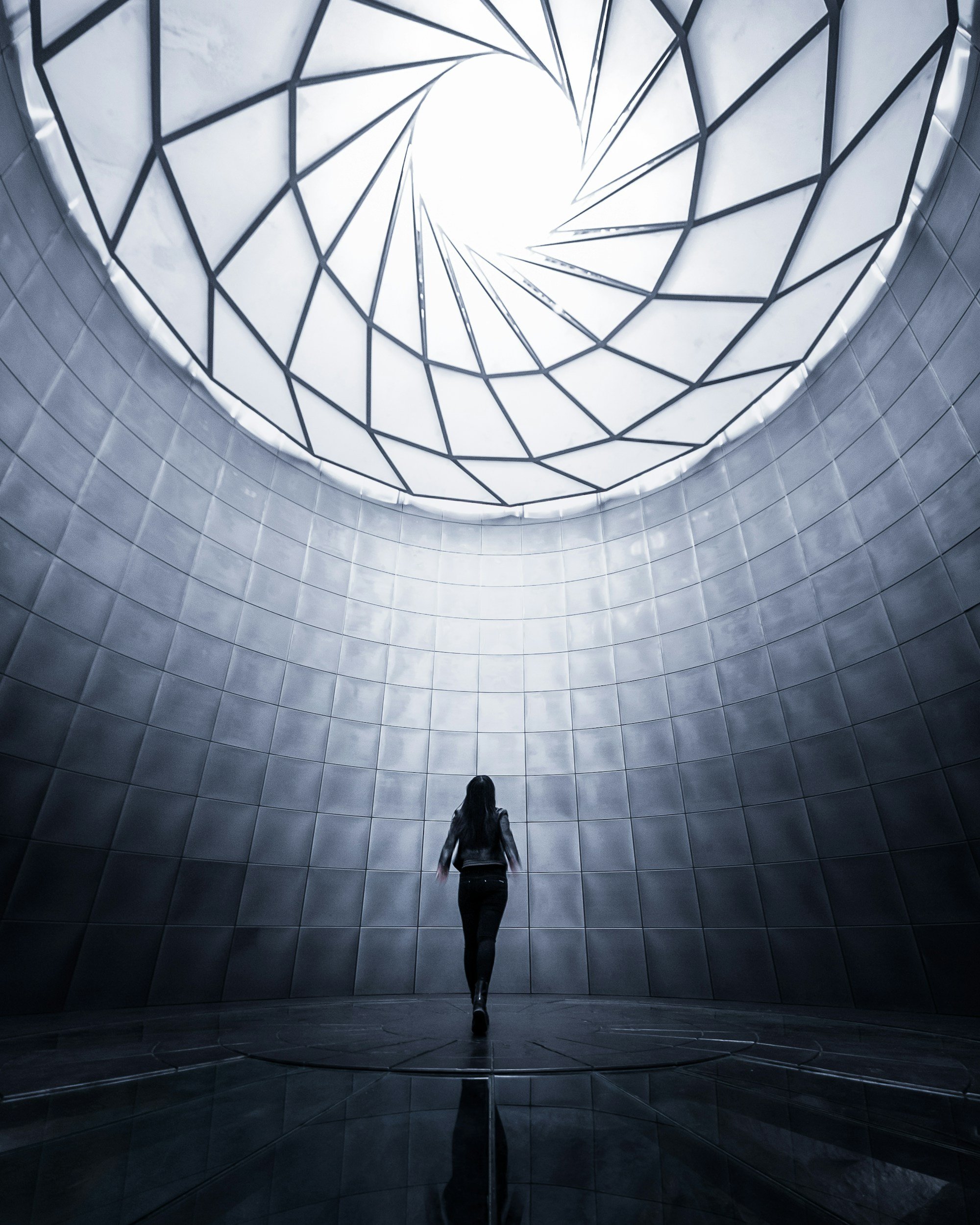 A woman walking inside a circular modern architectural space with a skylight ceiling featuring a geometric spiral design, and gray tiled walls.