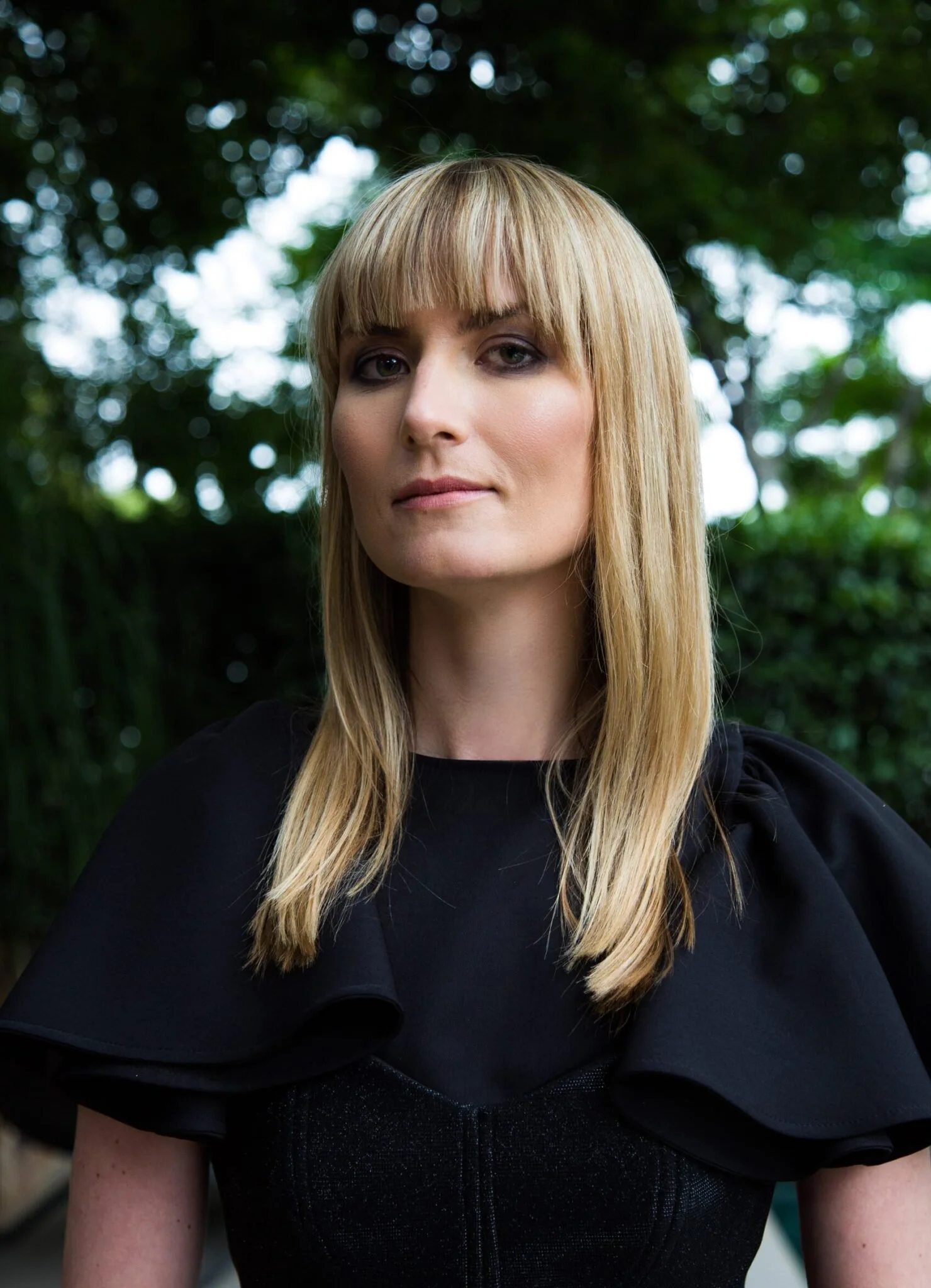 A woman with long blonde hair and bangs stands outdoors, wearing a black dress with puffed sleeves, with greenery and trees in the background.