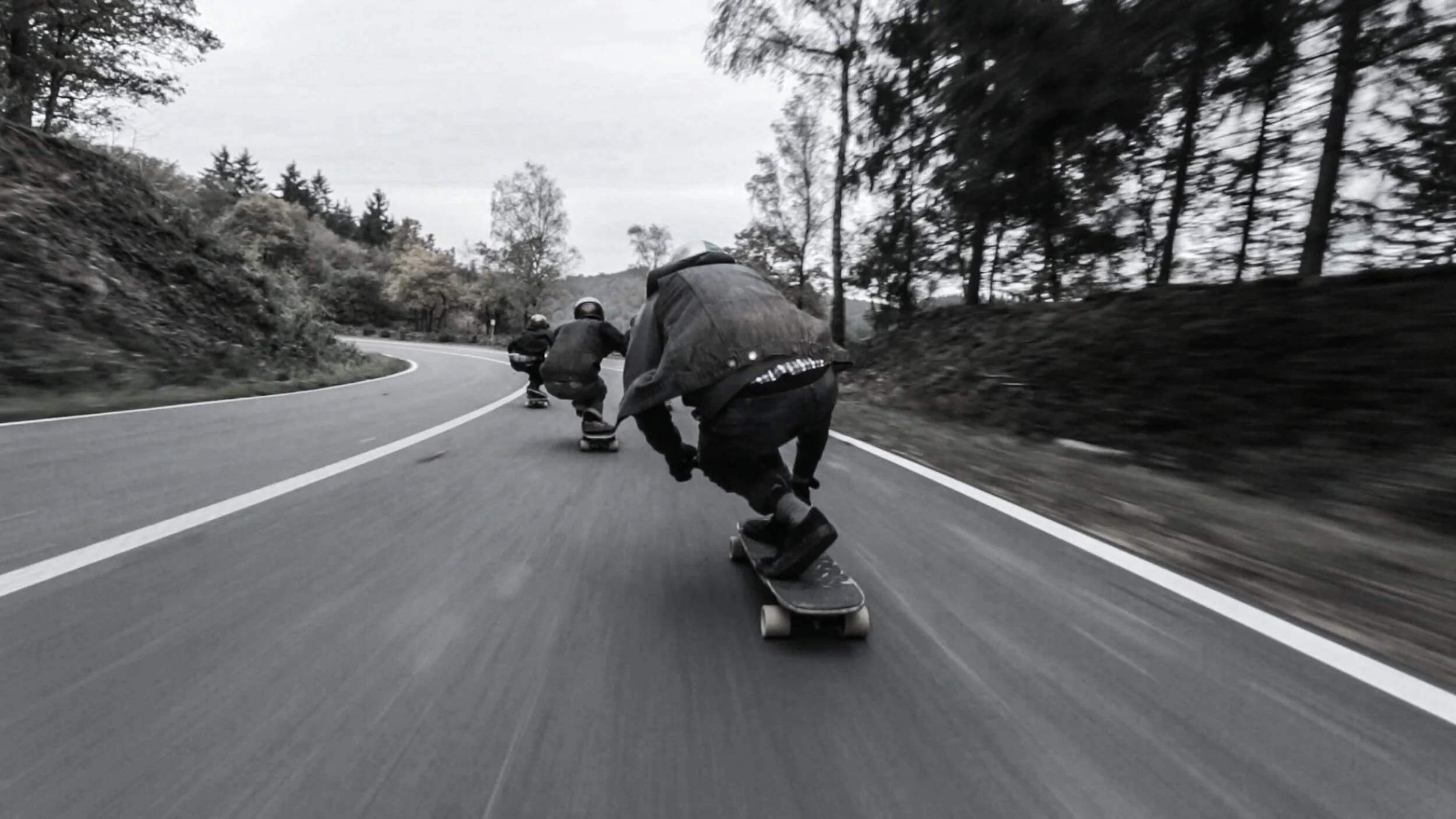 Three skateboarders riding downhill on a winding road through a wooded area.