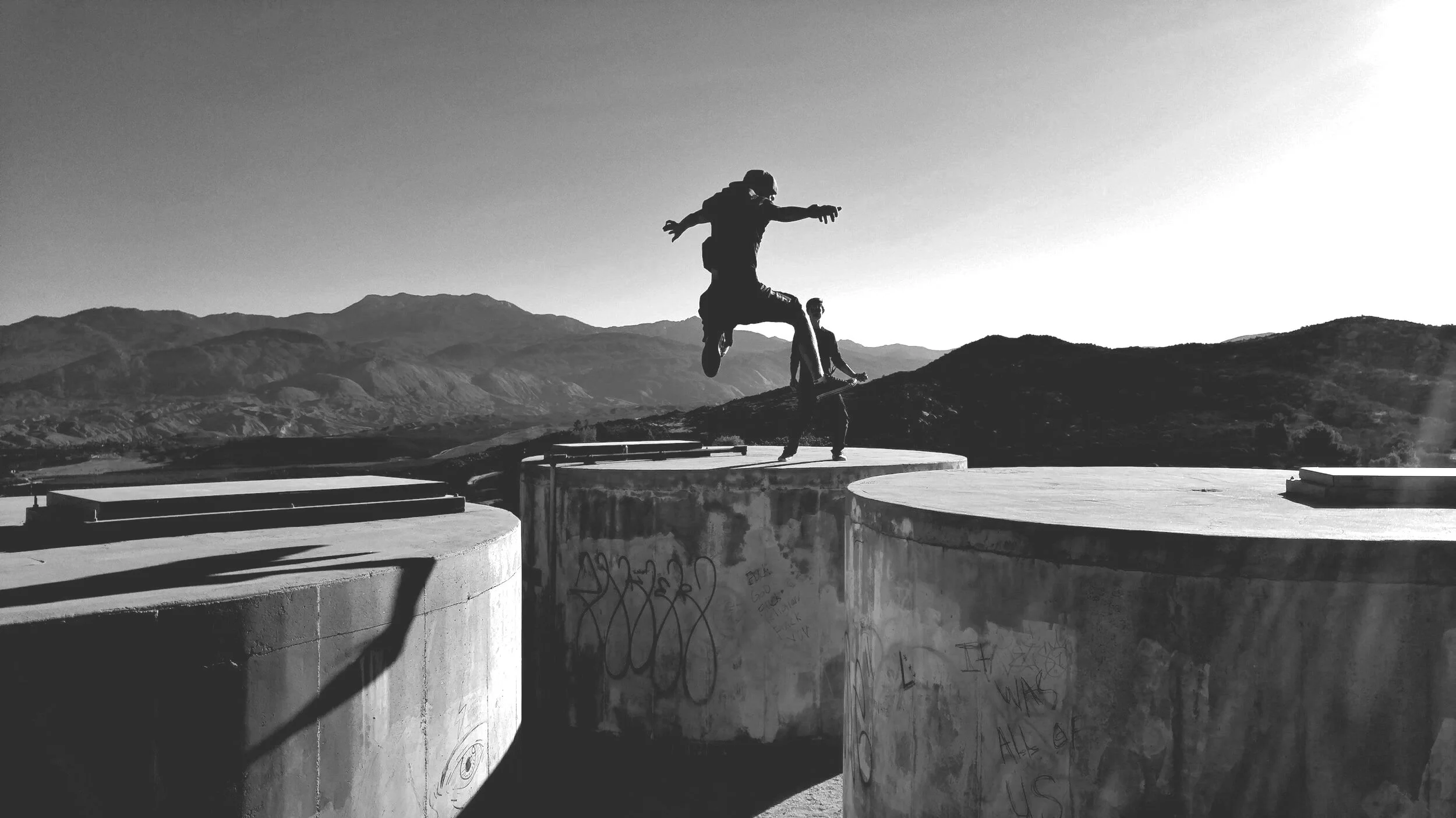 Two people on a rooftop performing parkour, one mid-air jump, with mountains in the background, black and white photo.