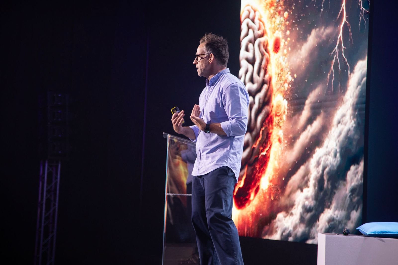 Man giving a presentation on stage with a large digital screen behind him displaying an image of a brain and an explosion, with clouds and lightning.
