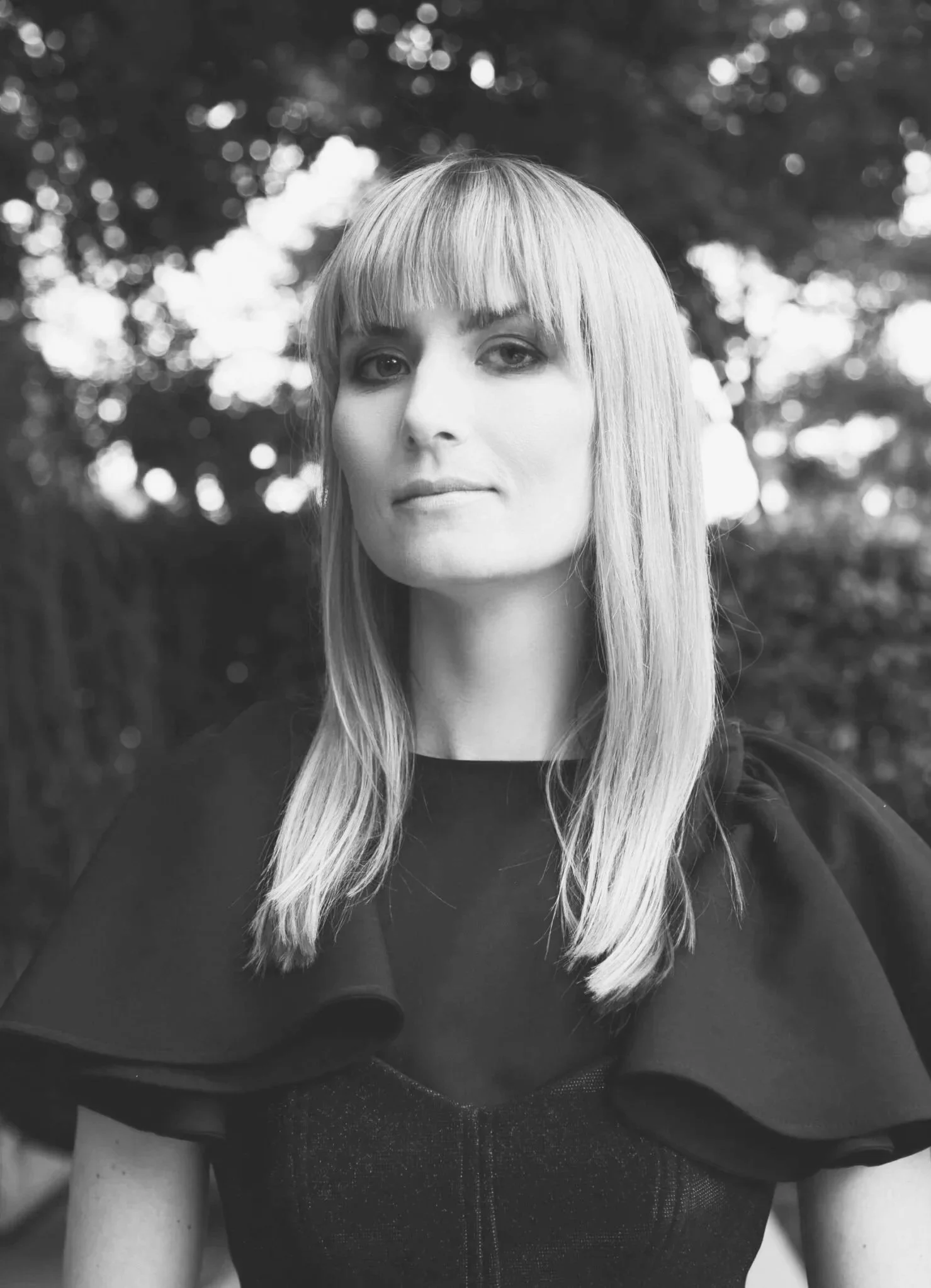 Black and white portrait of a woman with straight, shoulder-length hair and bangs, wearing a top with a structured shoulder design, outdoors with blurred trees in the background.