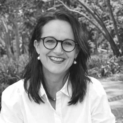 Black and white portrait of a woman with short dark hair, wearing glasses and a buttoned coat, standing outdoors in front of a blurred natural background.
