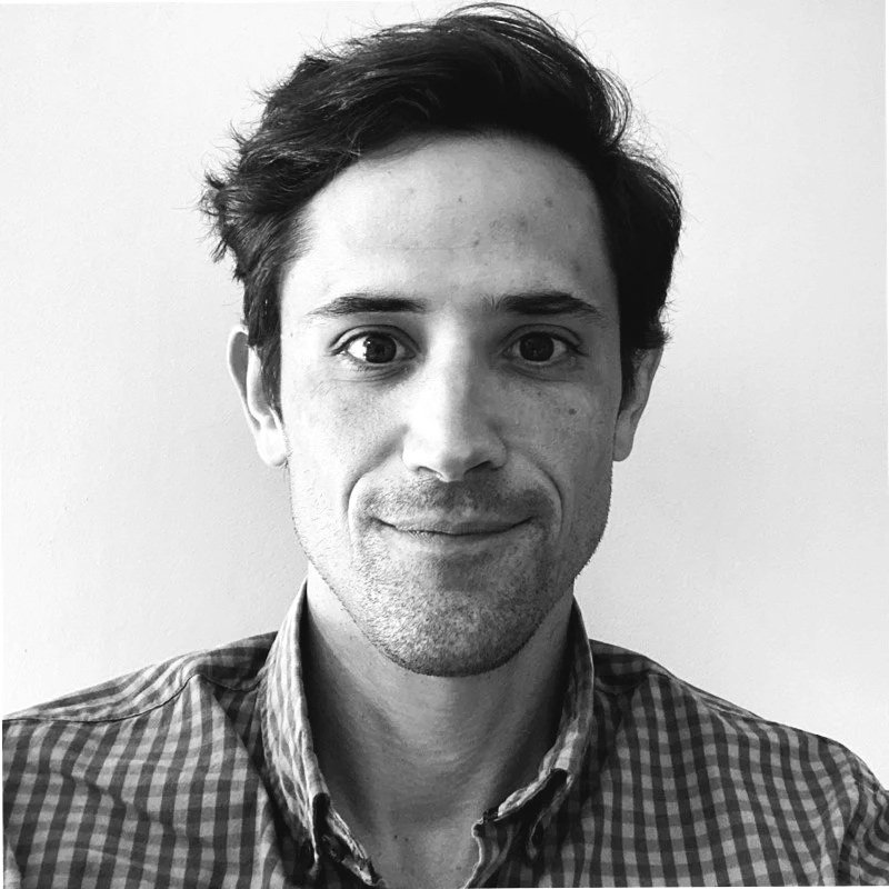 A black-and-white portrait of a man with short dark hair, wearing a plaid shirt, smiling slightly against a plain background.