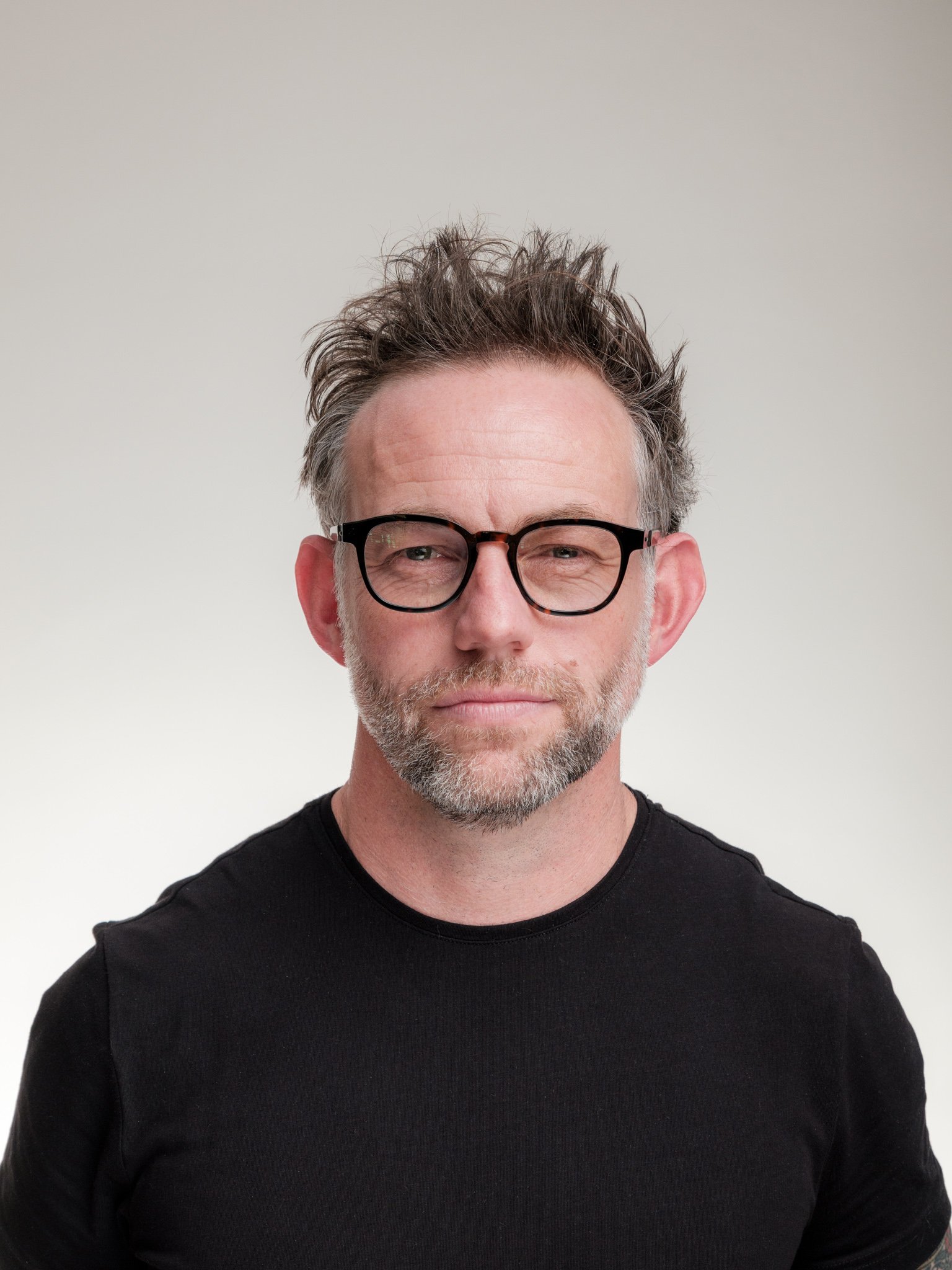 A middle-aged man with curly, grayish hair and a beard, wearing black-rimmed glasses and a black shirt, standing against a plain off-white background.