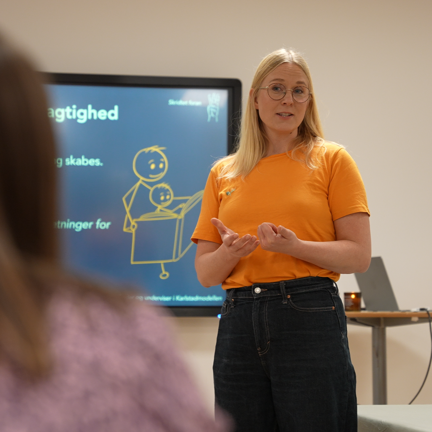 Kvinde i orange t-shirt taler foran en sort skærm med tekst i gul og hvid, omgivet af en illustration af en voksen og et barn, der læser sammen.