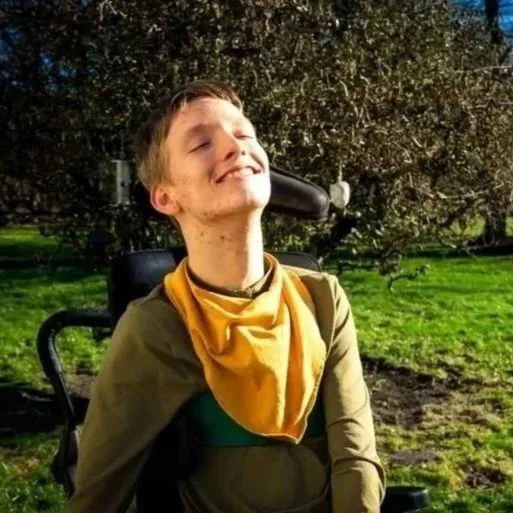 En person i kørestol nyder solen udendørs med grøn natur og træer i baggrunden.