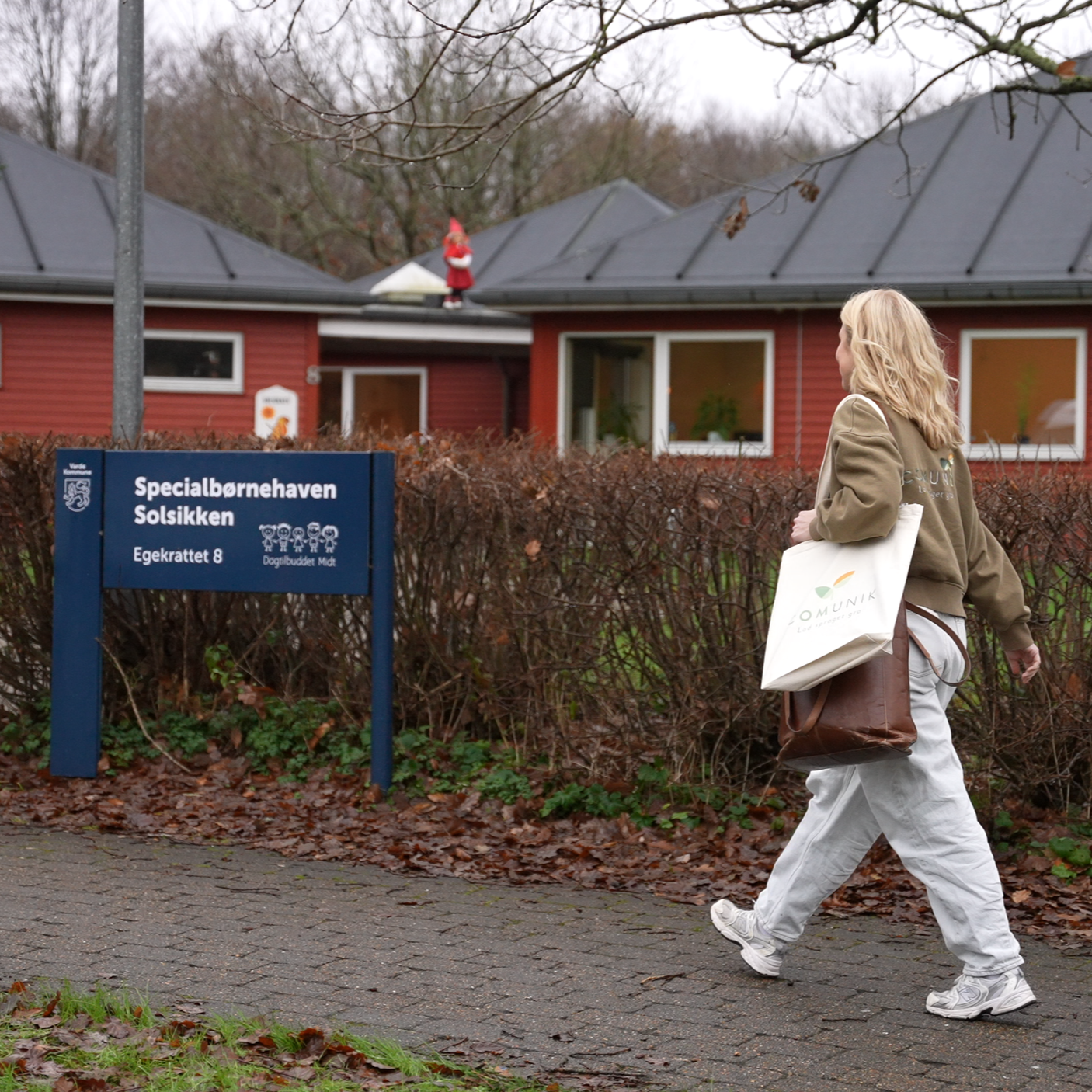 En kvinde går forbi indgangen til en børnehave kaldet 'Specialbørnehaven Solsikken' på Egekrattet 8, med et barn i baggrunden, der står på taget af en rød bygning.