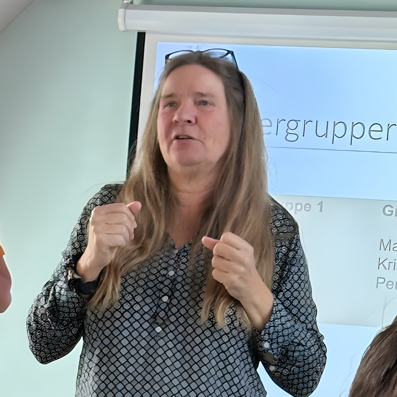 En kvinde med langt lyst hår, iført en sort og hvidt mønstret skjorte, står foran en whiteboard med en præsentation og ser ud til at tale.