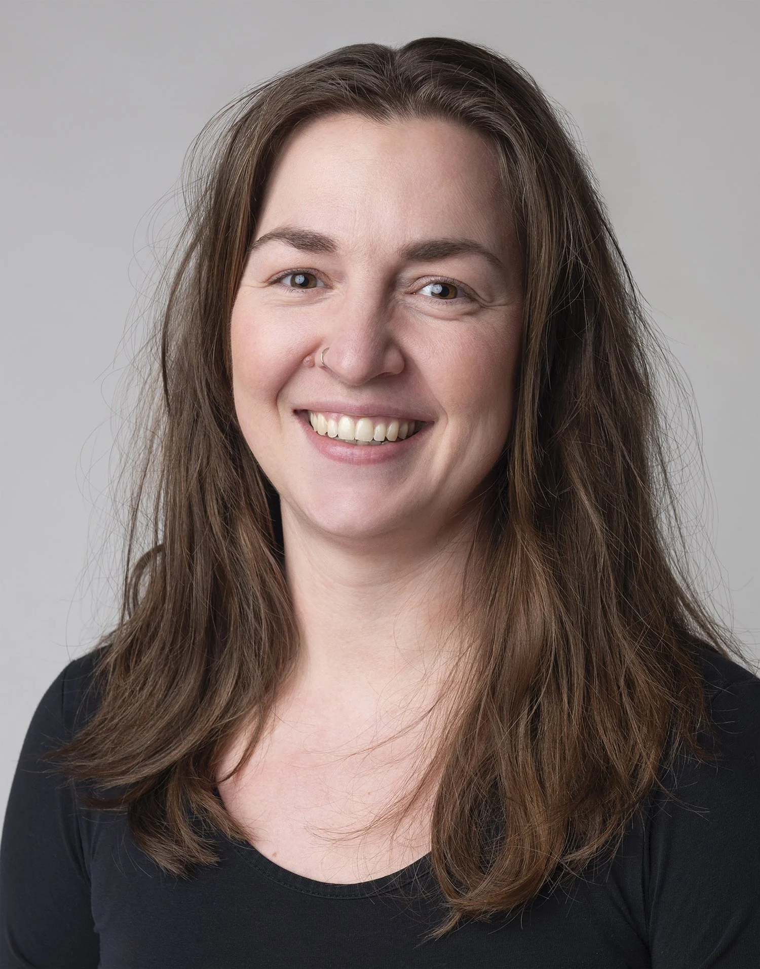 Smilende ung kvinde med mørkt hår og bruneøjne, iført sort tøj, står foran en neutral grå baggrund.