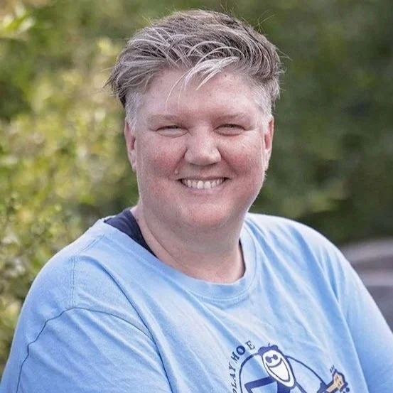 Person med kort, lyst hår smiler, iført lyseblåt T-shirt, udendørs baggrund med grønne blade.