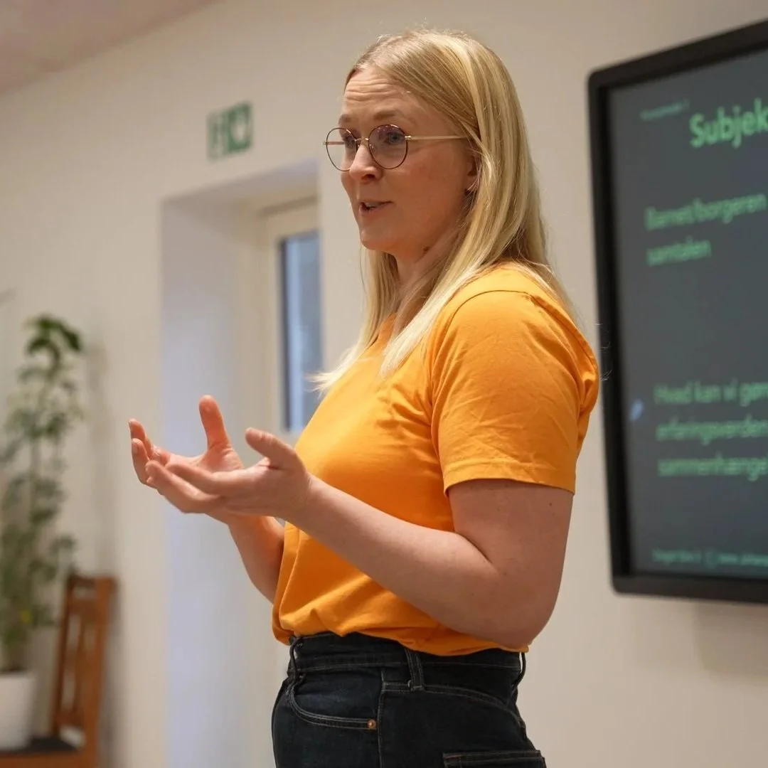 En kvinde med blondt hår, iført gult t-shirt og sorte jeans, står foran en skærm og taler, mens hun bruger håndbevægelser.